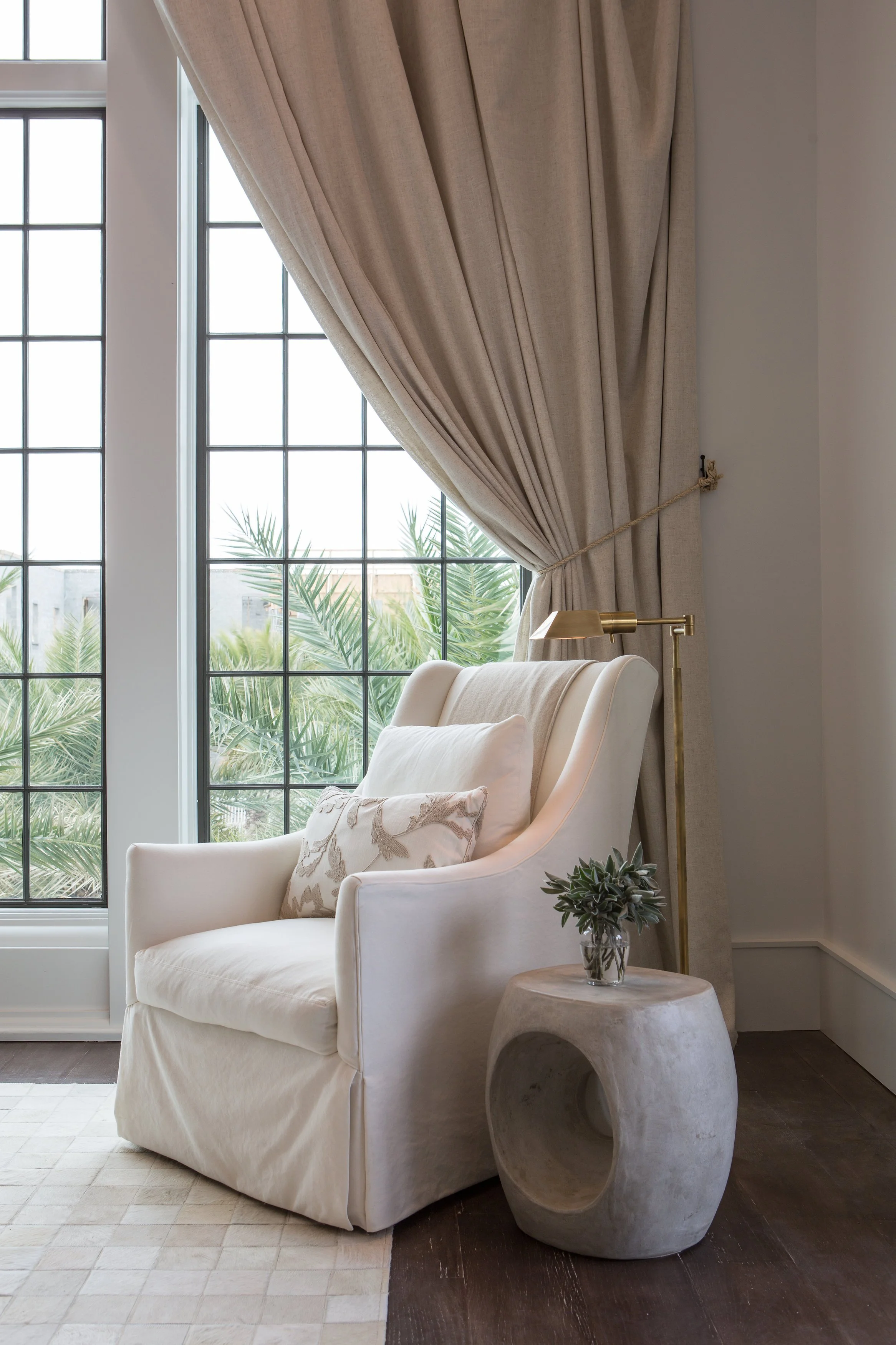 A cozy corner with a white armchair, decorative pillows, a small concrete side table with a plant, and floor-to-ceiling beige curtains near a large window showing greenery outside.