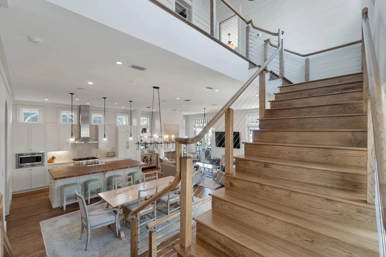 Open-concept living space with kitchen, dining, and living areas. The kitchen has white cabinets, a wooden island, and stainless steel appliances. There is a chandelier above the dining table, and a staircase with wooden steps and a railing leading u