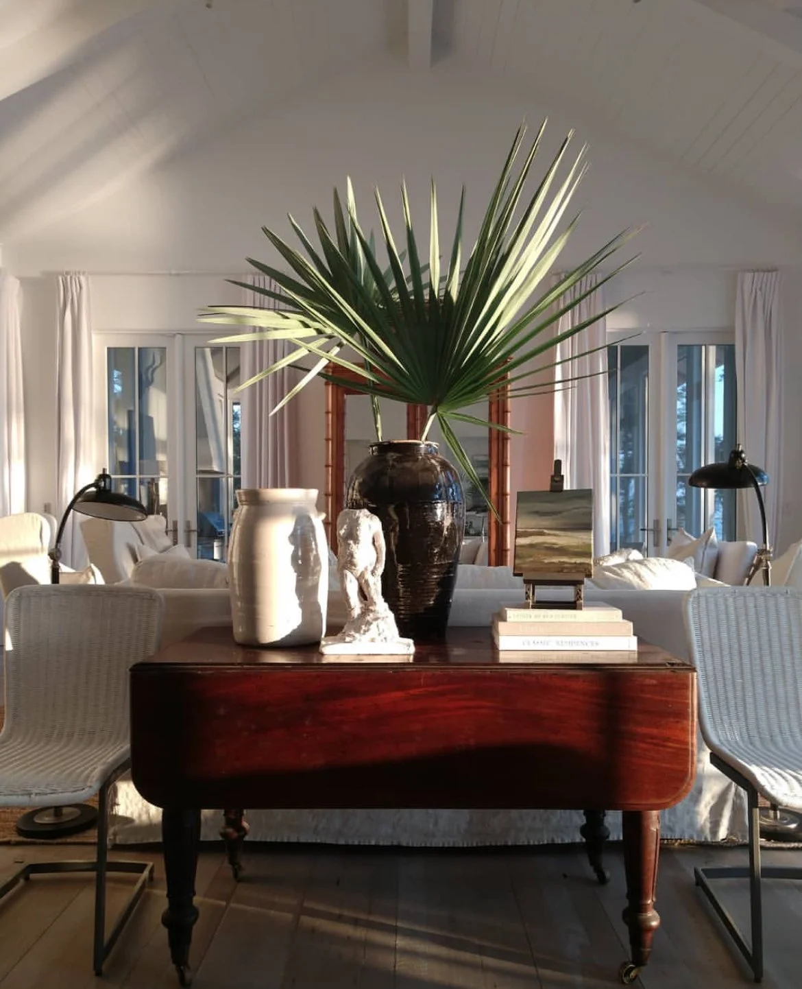 A living room with a wooden table holding a large potted palm leaf, white vases, a small sculpture, and stacked books, with chairs and a sofa in the background.