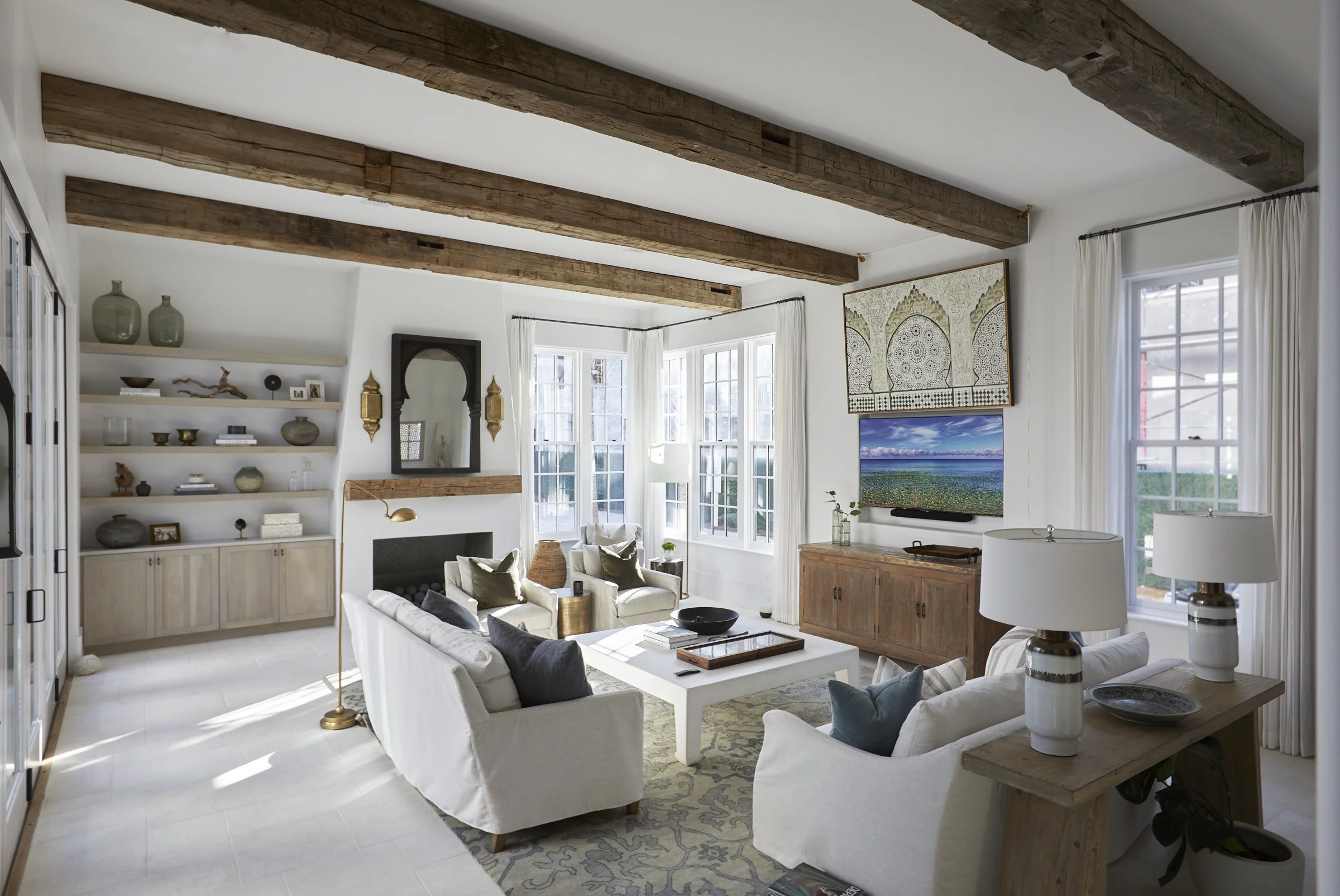 Bright living room with white sofas, wooden beams on ceiling, built-in shelves with decor, large windows with white curtains, fireplace, wall-mounted TV, and a wooden console table.