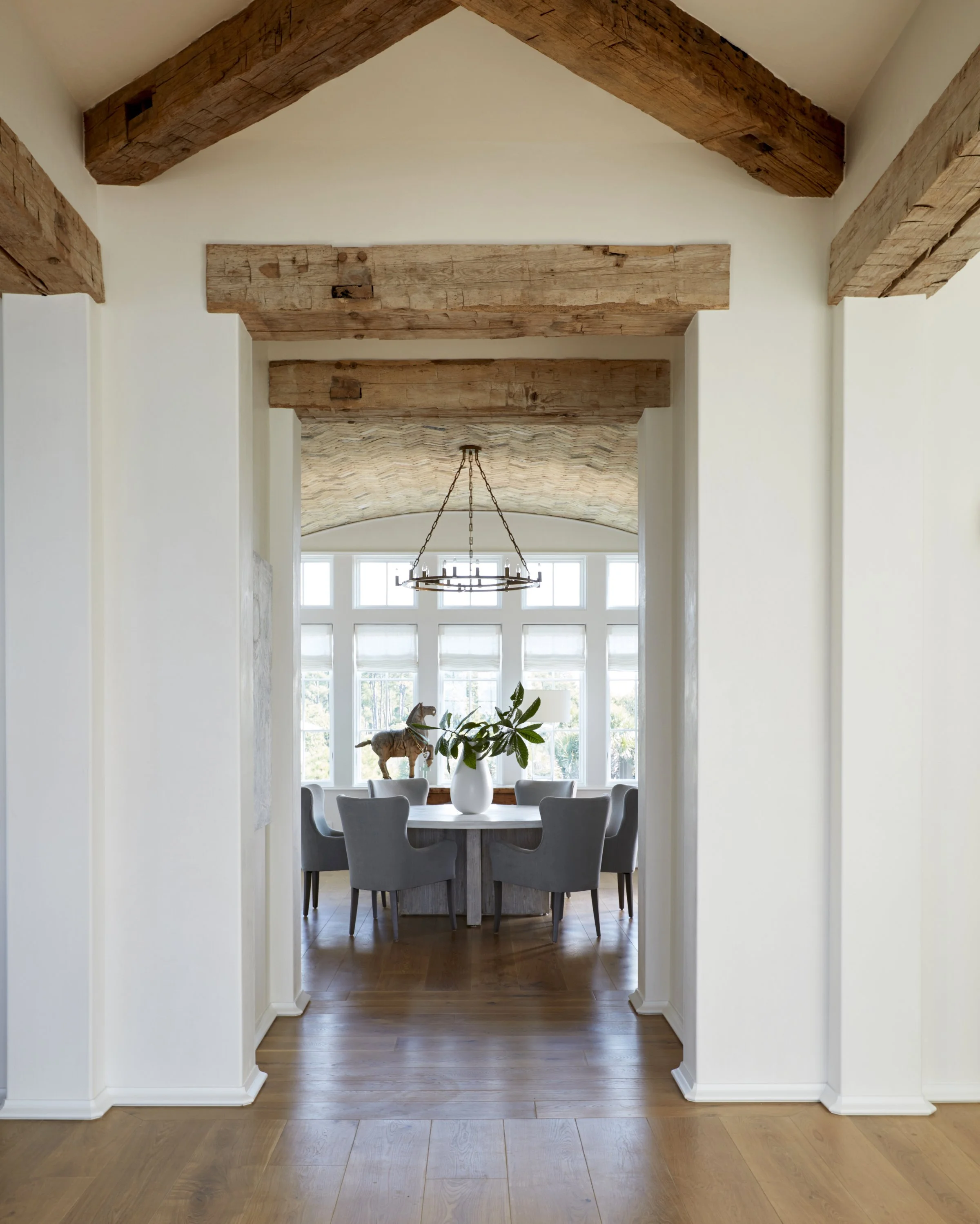 View through a hallway into a dining room with large windows, a round table, six gray chairs, a white vase with green leaves, and a throw horse on the windowsill, with wooden beams and a chandelier overhead.