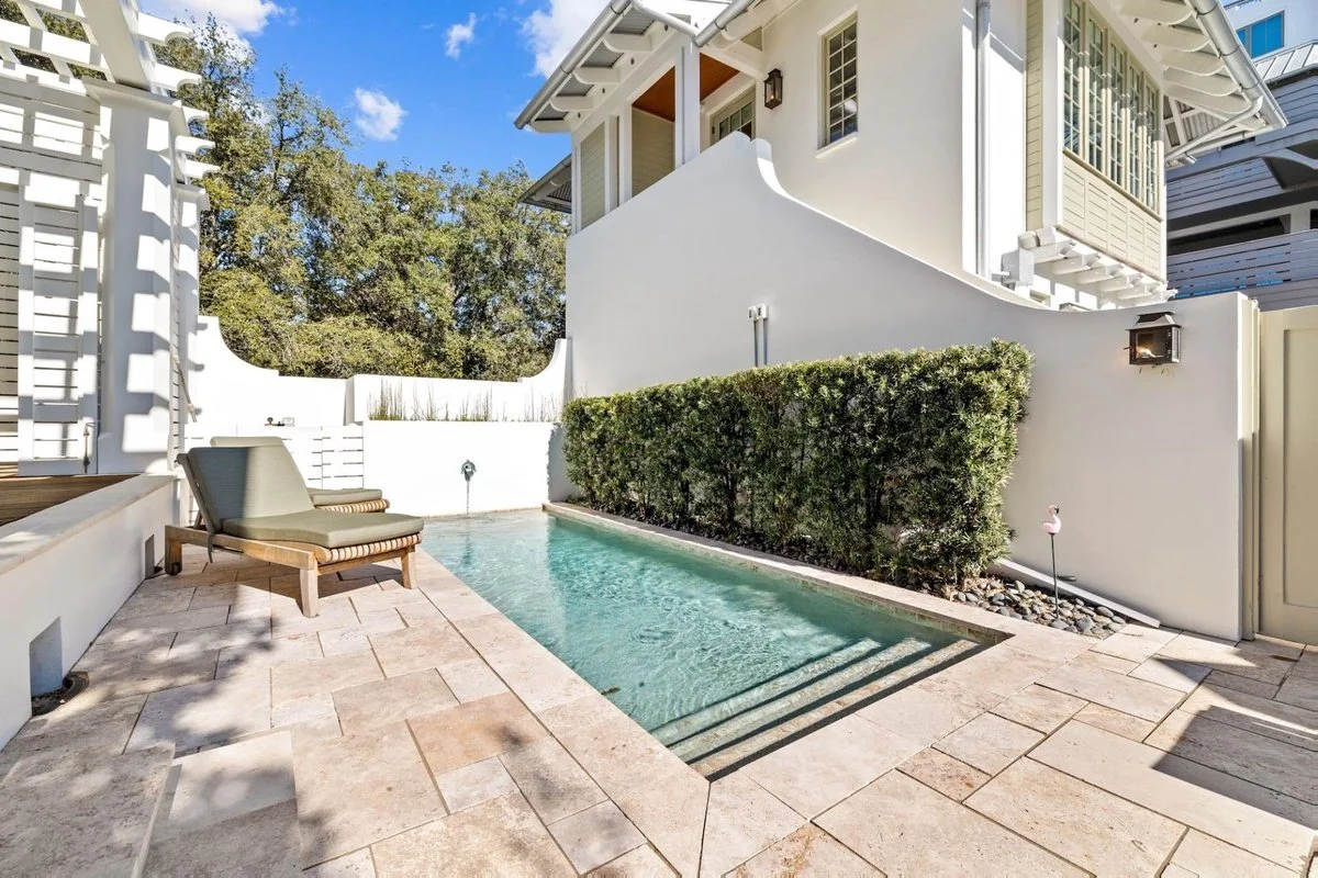Small rectangular swimming pool with steps at one end, neighboring a tiled patio area with lounge chairs, enclosed by white walls and greenery, adjacent to a multi-story house with light-colored exterior and windows, under a blue sky with a few cloud