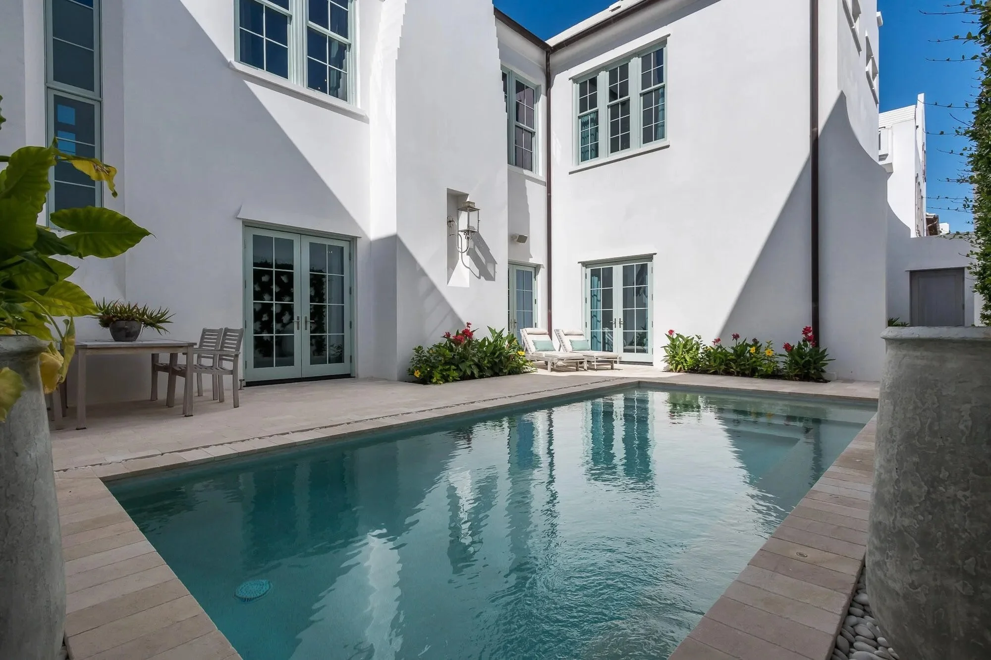 View of a backyard with a small swimming pool, white house walls, patio furniture, and potted plants, under a clear blue sky.