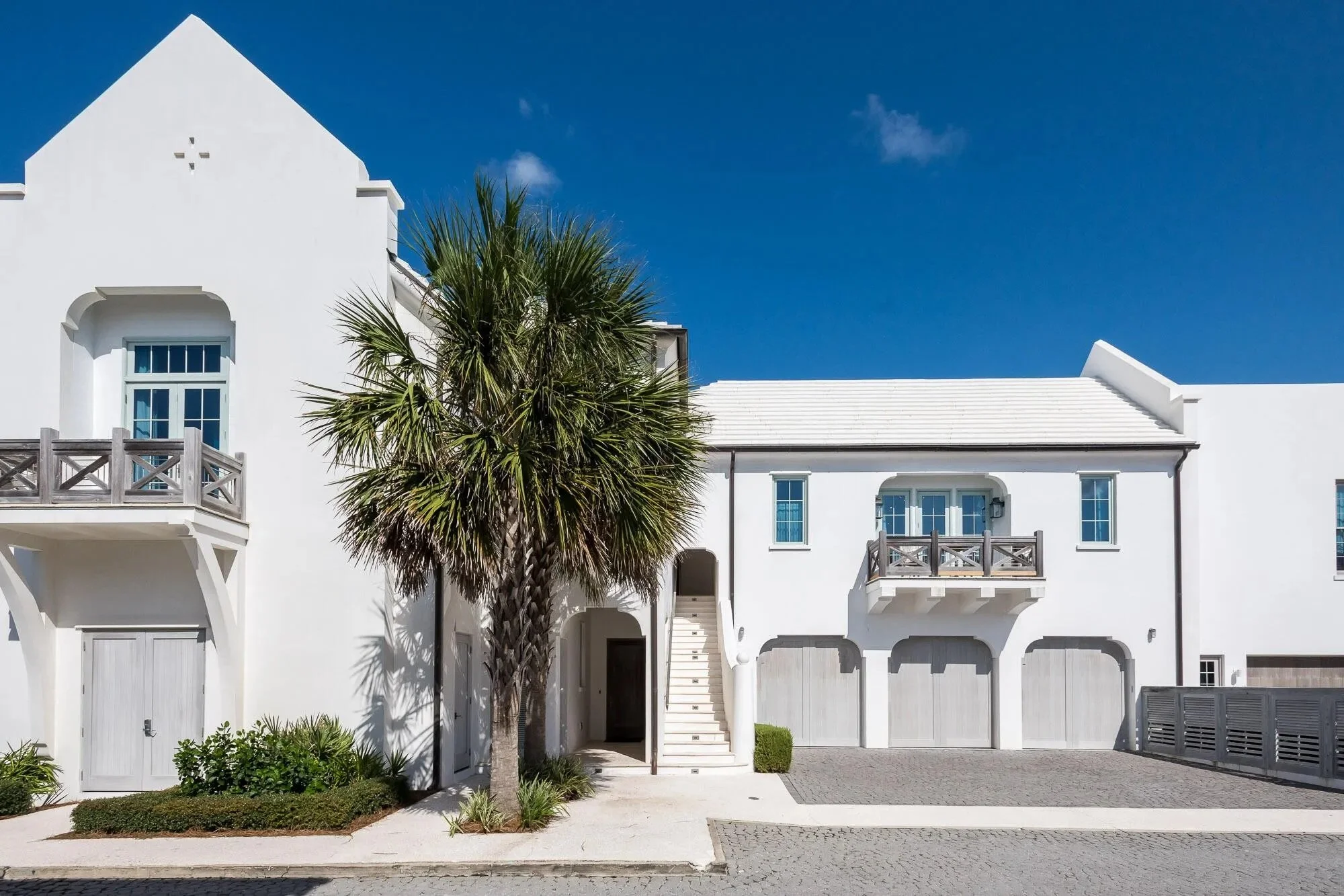 A white modern house with a staircase, small balconies, and a palm tree in front, under a clear blue sky.
