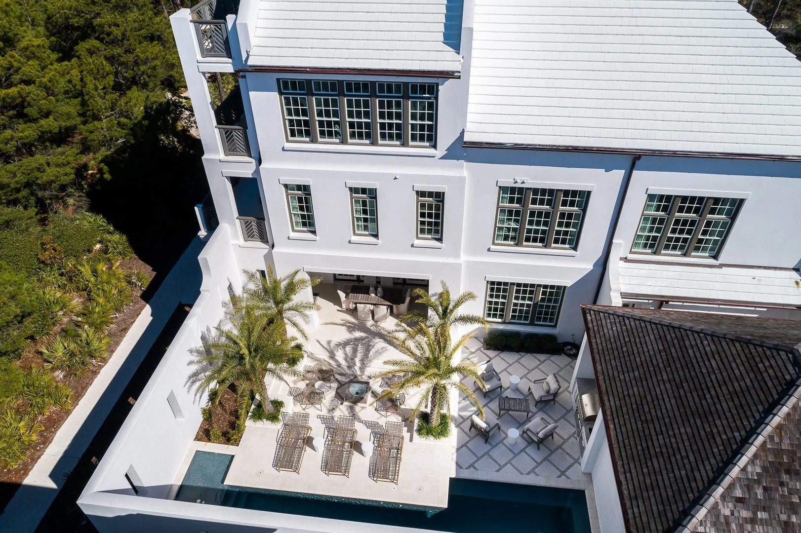 Aerial view of a white multi-story house with large windows, a patio with outdoor furniture, palm trees, and a landscaped yard surrounded by lush greenery.