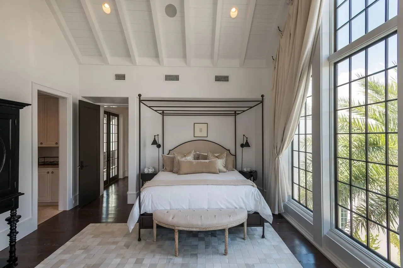 A bedroom with a four-poster bed, beige and white bedding, black side tables with lamps, large windows with curtains, and a small cushioned bench at the foot of the bed.
