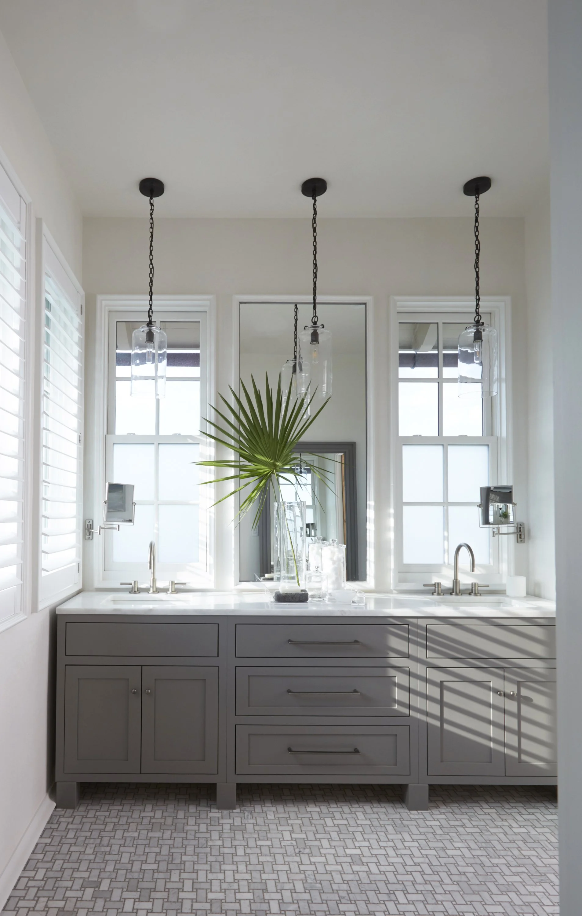Bright bathroom with gray vanity, pendant lights, and large windows with white shutters.