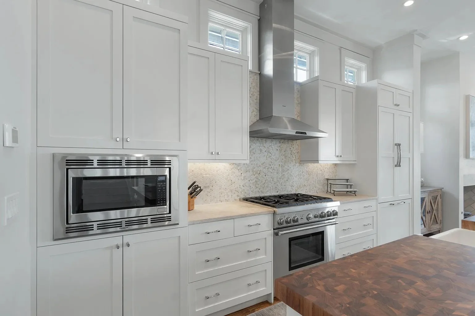 Modern kitchen with white cabinets, stainless steel microwave, stove, and range hood, wooden table, and small window above the countertop.