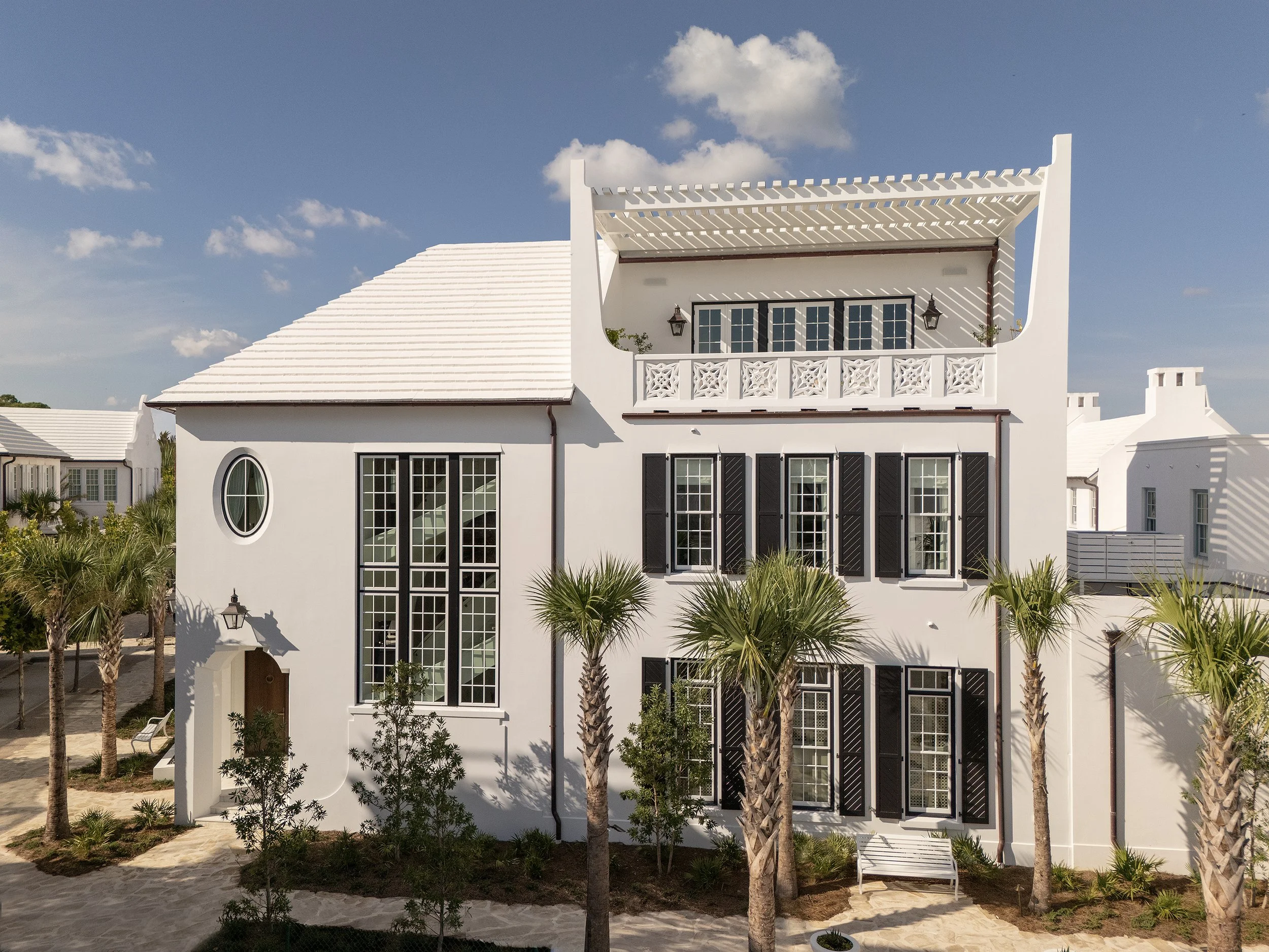 White multi-story house with black shutters, palm trees in front, and a sand pathway, under a partly cloudy sky.