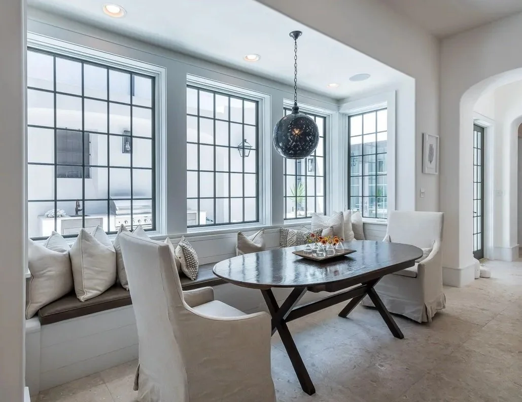 A bright dining area with large windows, a dark oval wooden table, white upholstered chairs, a built-in window seat with pillows, and a black ceiling pendant light.