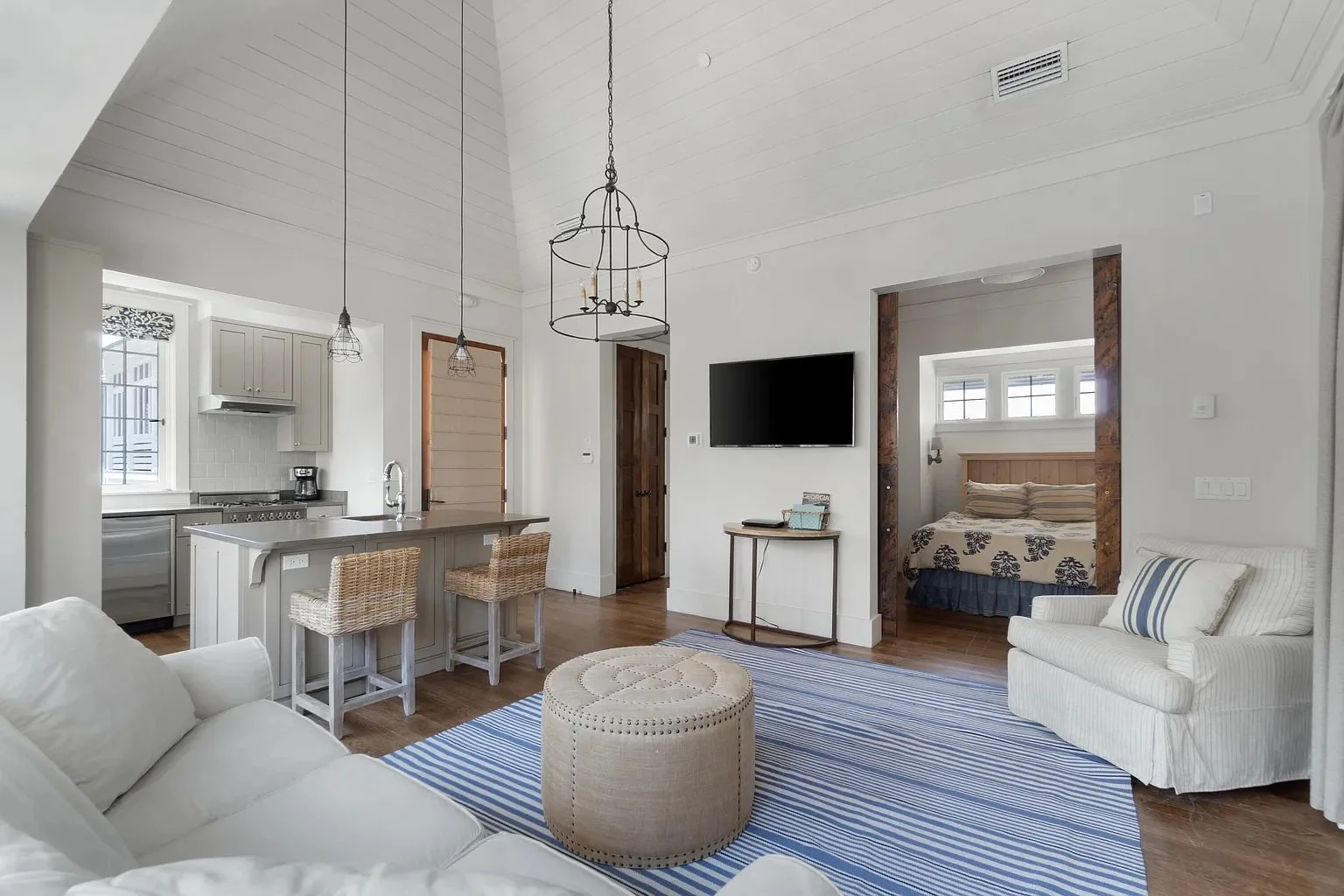 Living room with white walls, wooden floors, and a high ceiling. Contains a white sofa, a striped armchair, a round beige ottoman, a wall-mounted TV, a small table, and a bedroom doorway. Kitchen visible with gray cabinetry and a breakfast bar with w
