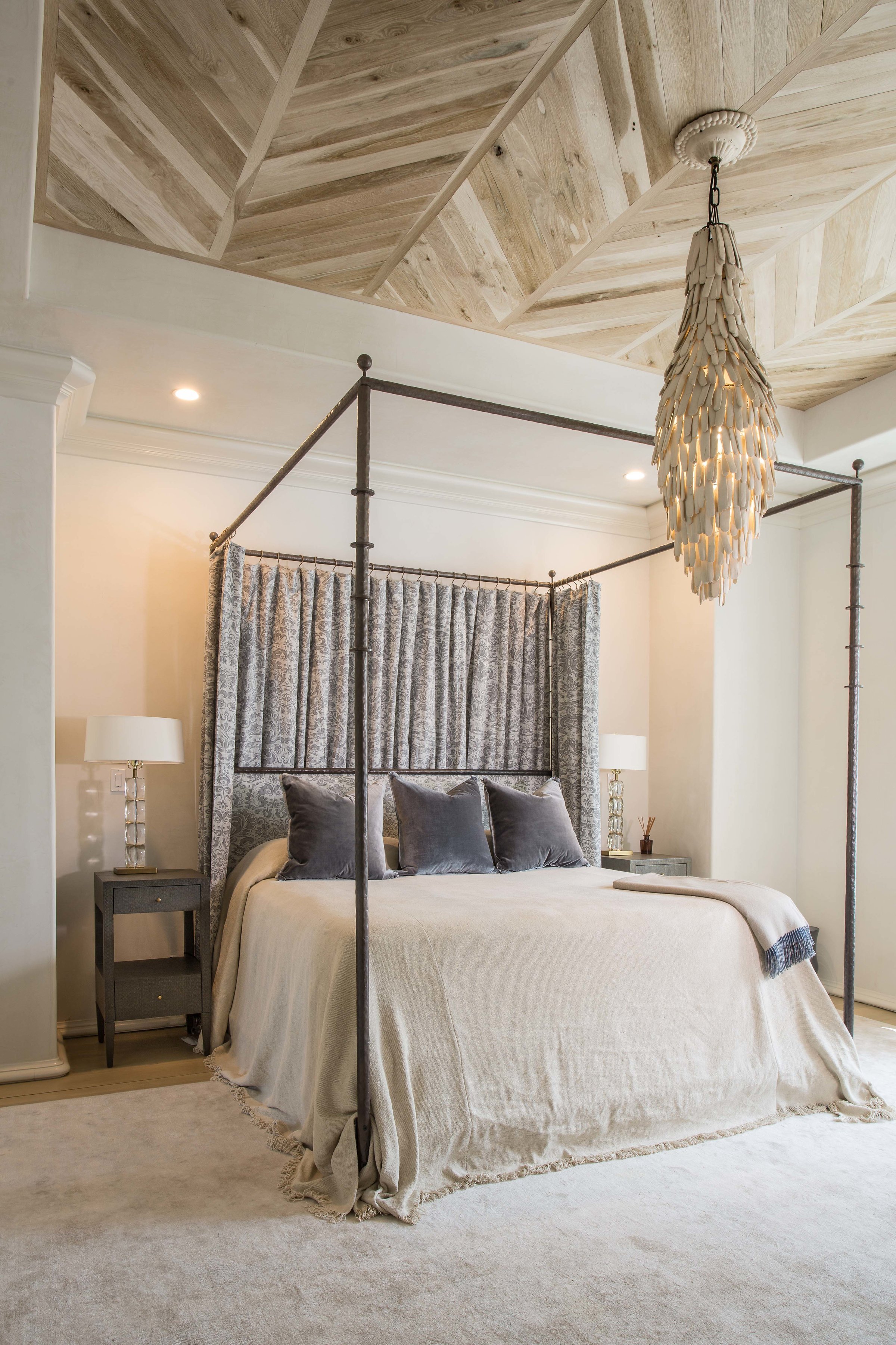 A bedroom with a beige bed, gray pillows, two matching nightstands with lamps, and a curtain behind the bed. A textile chandelier hangs from the ceiling, and the ceiling has a wood panel design.