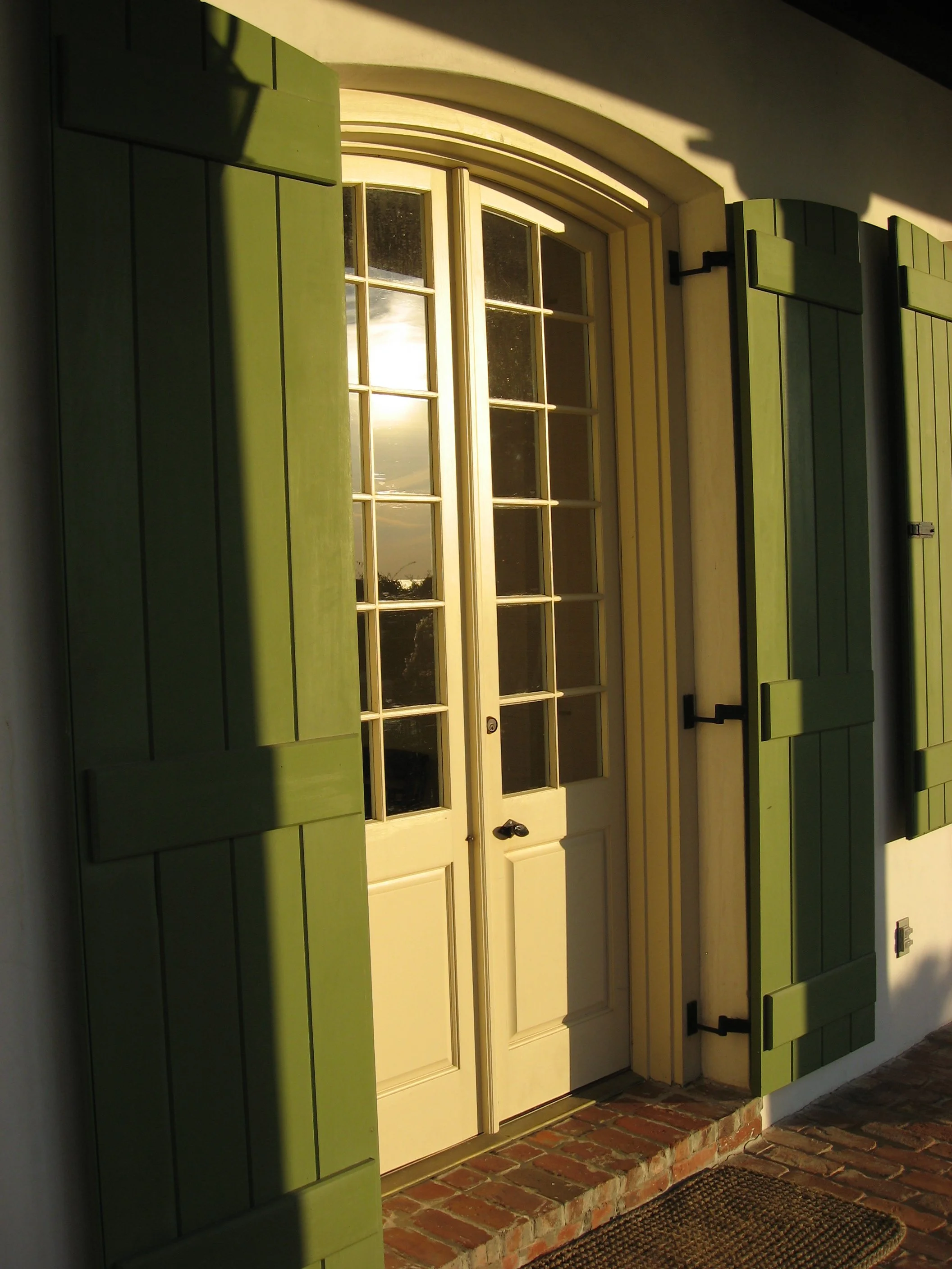 A white door with glass panels, surrounded by green shutters, and positioned on a brick porch, with sunlight casting shadows.