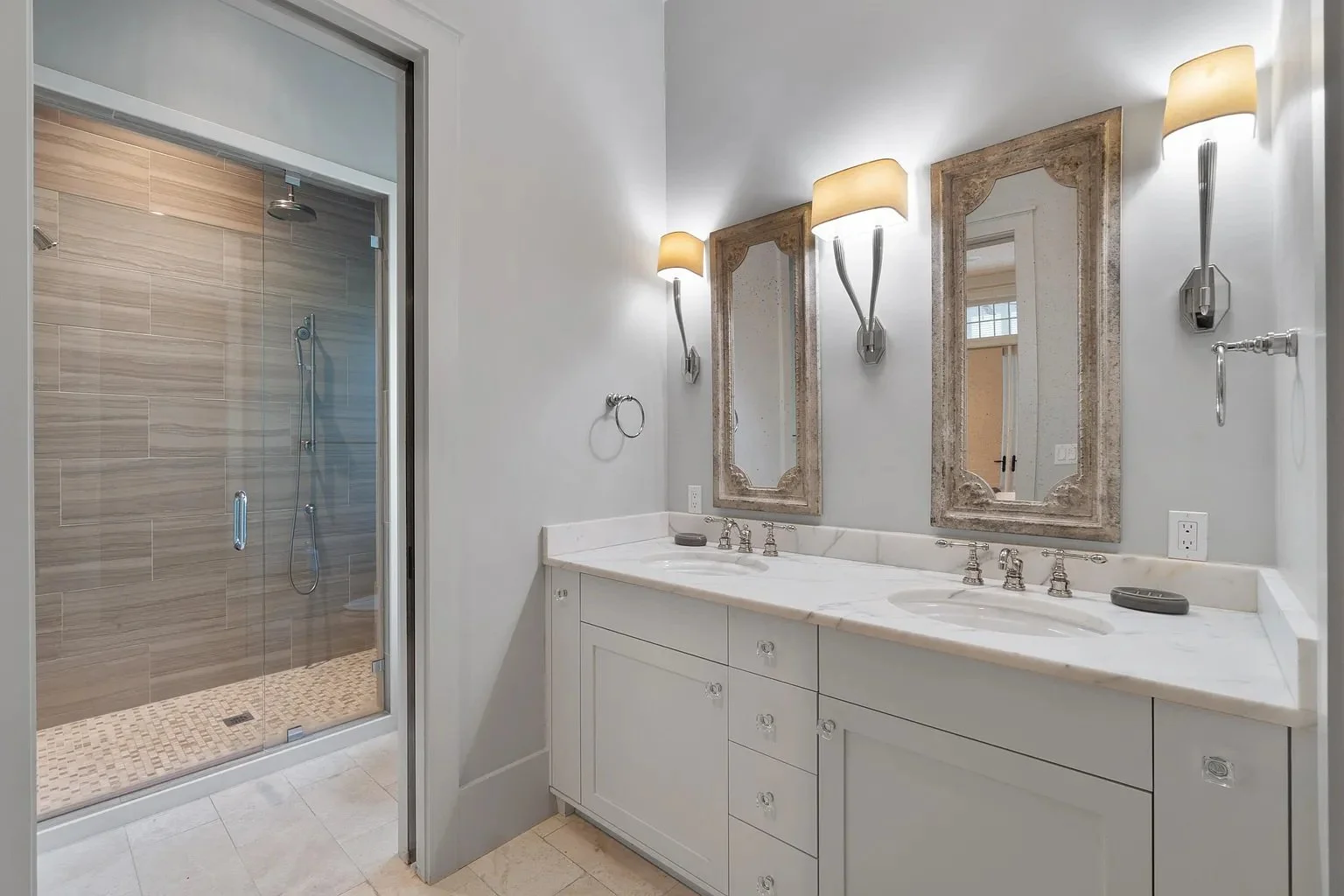 Bathroom with a double sink vanity, two framed mirrors, and decorative wall sconces; a walk-in shower with a glass door and beige tiles is visible to the left.