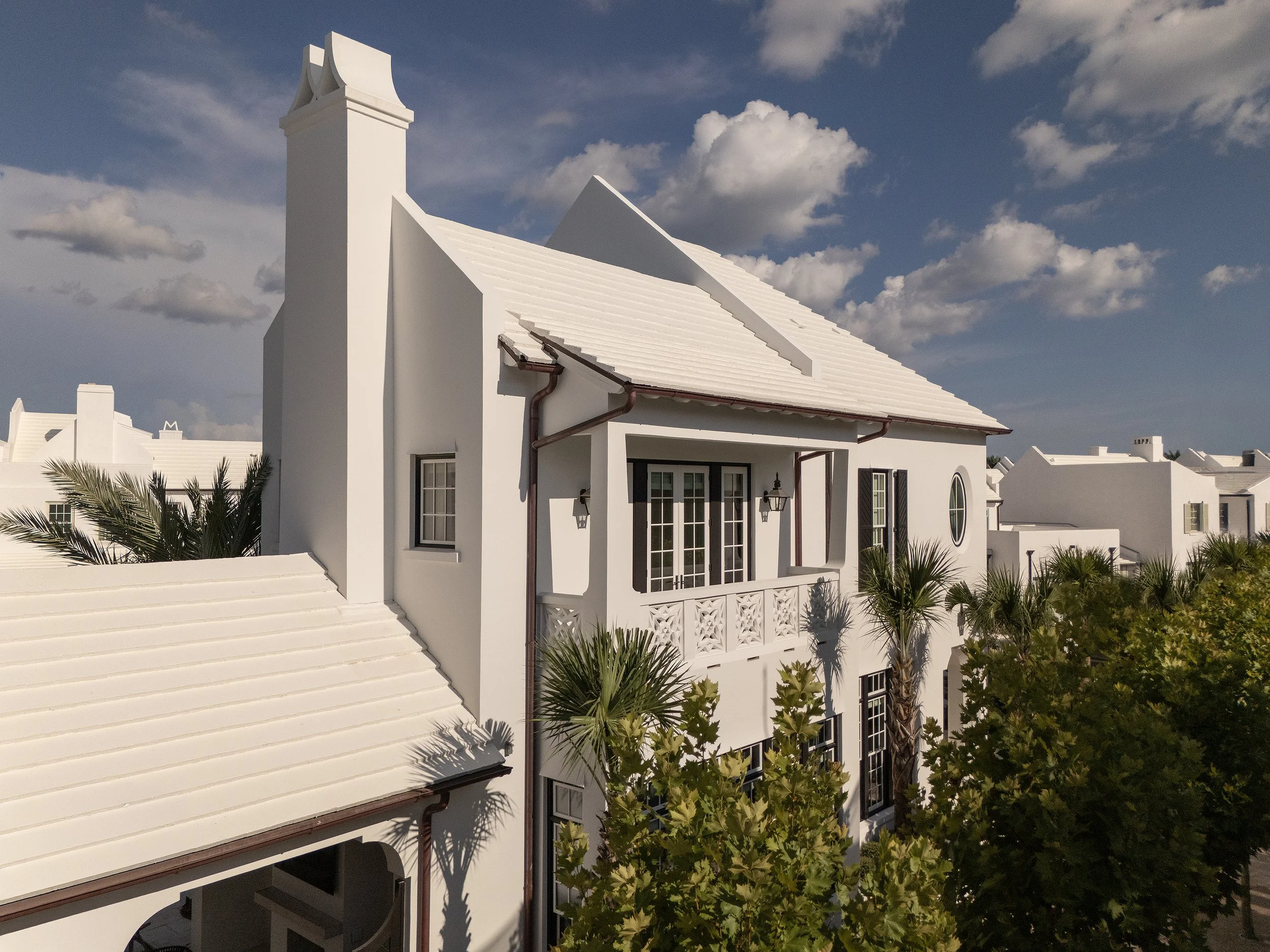 White house with a steep roof, multiple windows, and a small balcony, surrounded by palm trees and other greenery, under a partly cloudy sky.