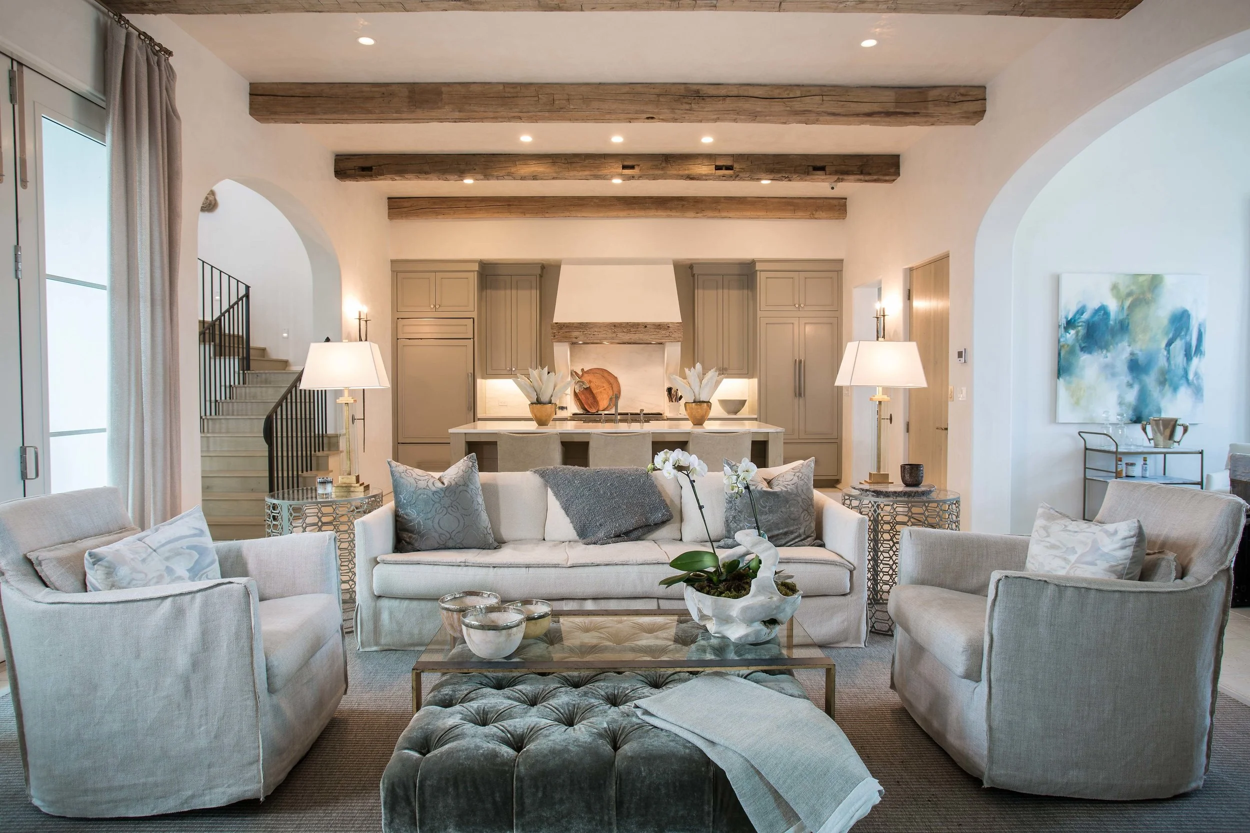 Living room with a white sofa, two armchairs, a glass coffee table, and a gray tufted ottoman, decorated with pillows and a orchid plant. In the background, open kitchen with beige cabinets, a white island, and decorative vases, with ceiling beams an