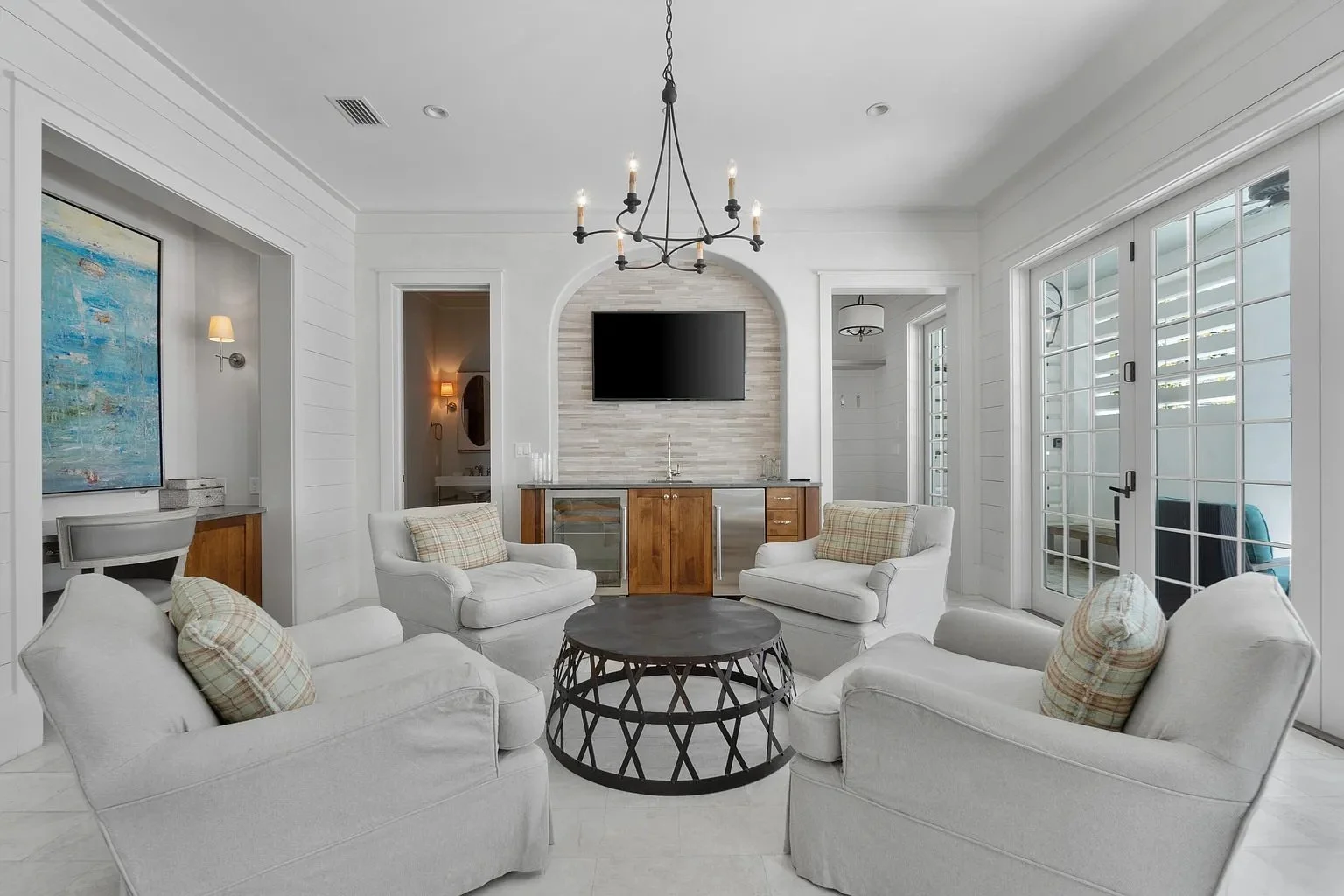 A bright living room with white walls, ceiling, and furniture, featuring four white armchairs with plaid pillows around a dark round coffee table, a wall-mounted flat-screen TV, a chandelier, and large glass doors leading outside.