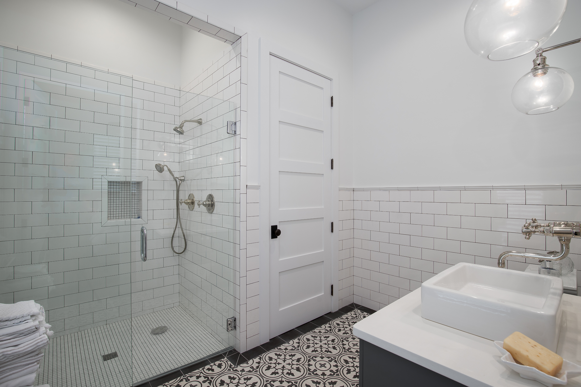 Modern bathroom with a walk-in glass shower, white subway tile walls, a black and white patterned floor, a white vanity with a vessel sink, and a soap bar on a dish.