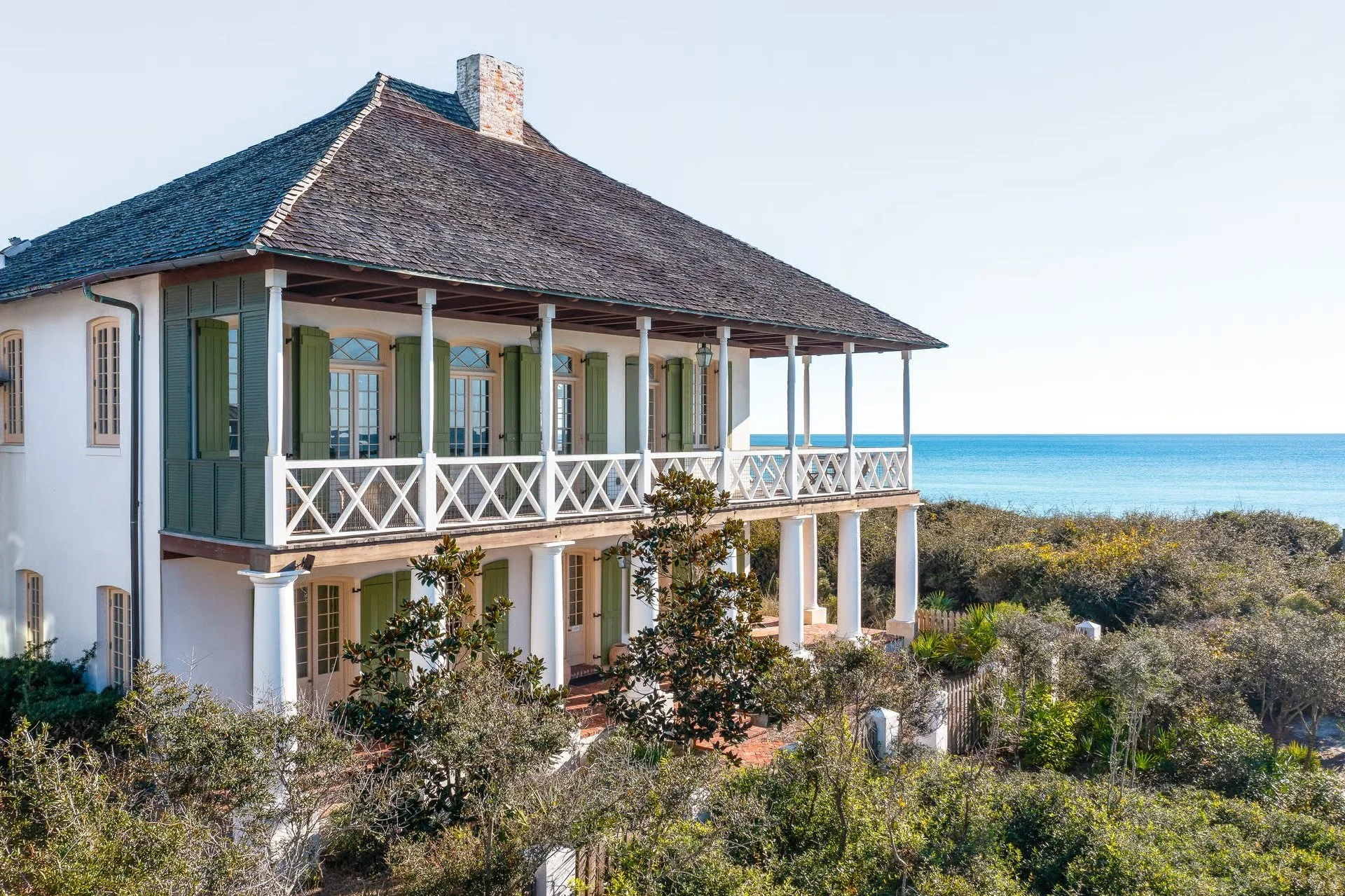 A large seaside house with a balcony, green shutters, and a tiled roof surrounded by greenery, overlooking the ocean.