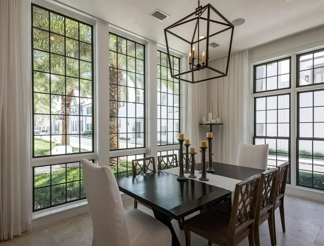 Dining room with large windows, a black dining table set with candles, white and wooden chairs, a geometric candle holder hanging from the ceiling, and white curtains.