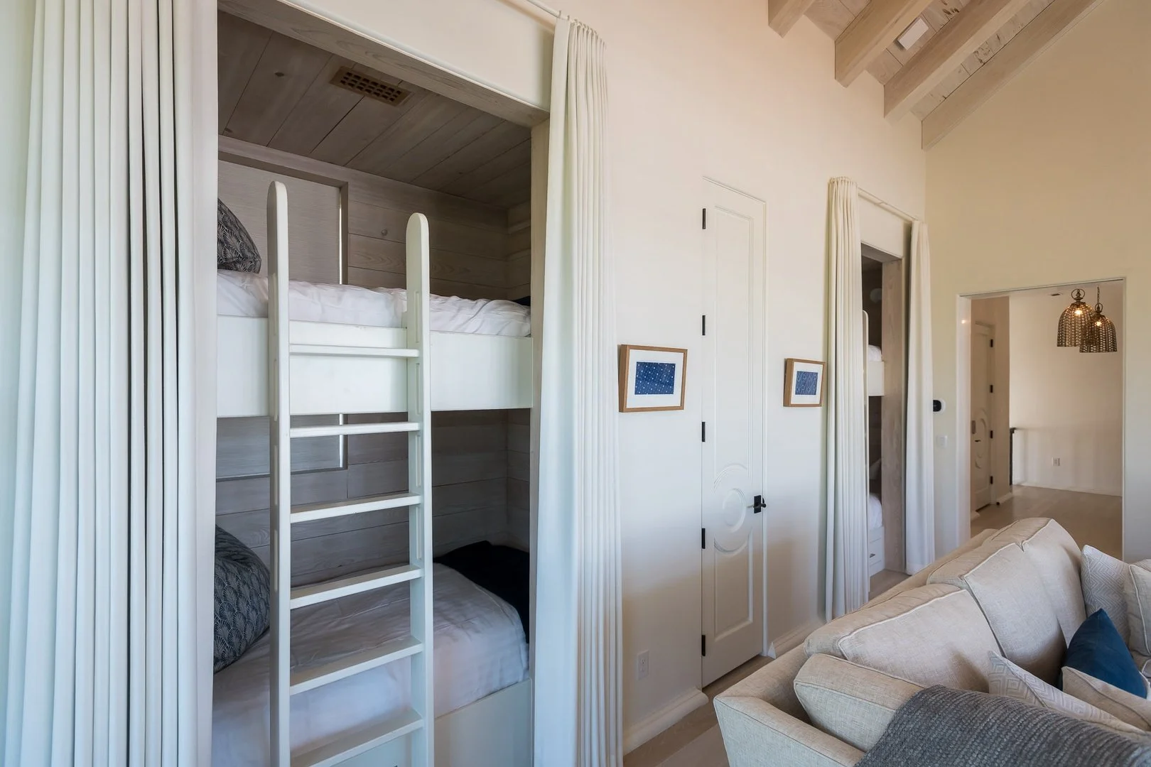 Interior of a living space with a built-in bunk bed, white curtains, a beige sofa with pillows, framed artwork on the wall, and a doorway leading to another room with a hanging light fixture.