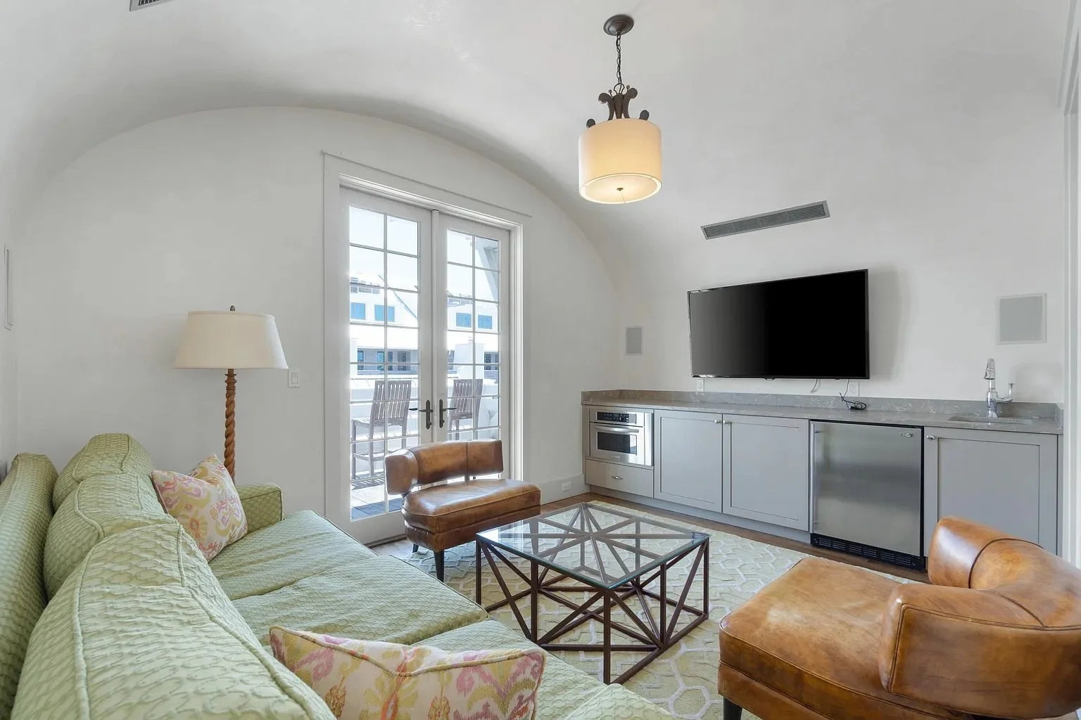 Living room with glass door leading to balcony, furnished with a green sofa, brown leather chairs, a glass coffee table, and a wall-mounted flat-screen TV.