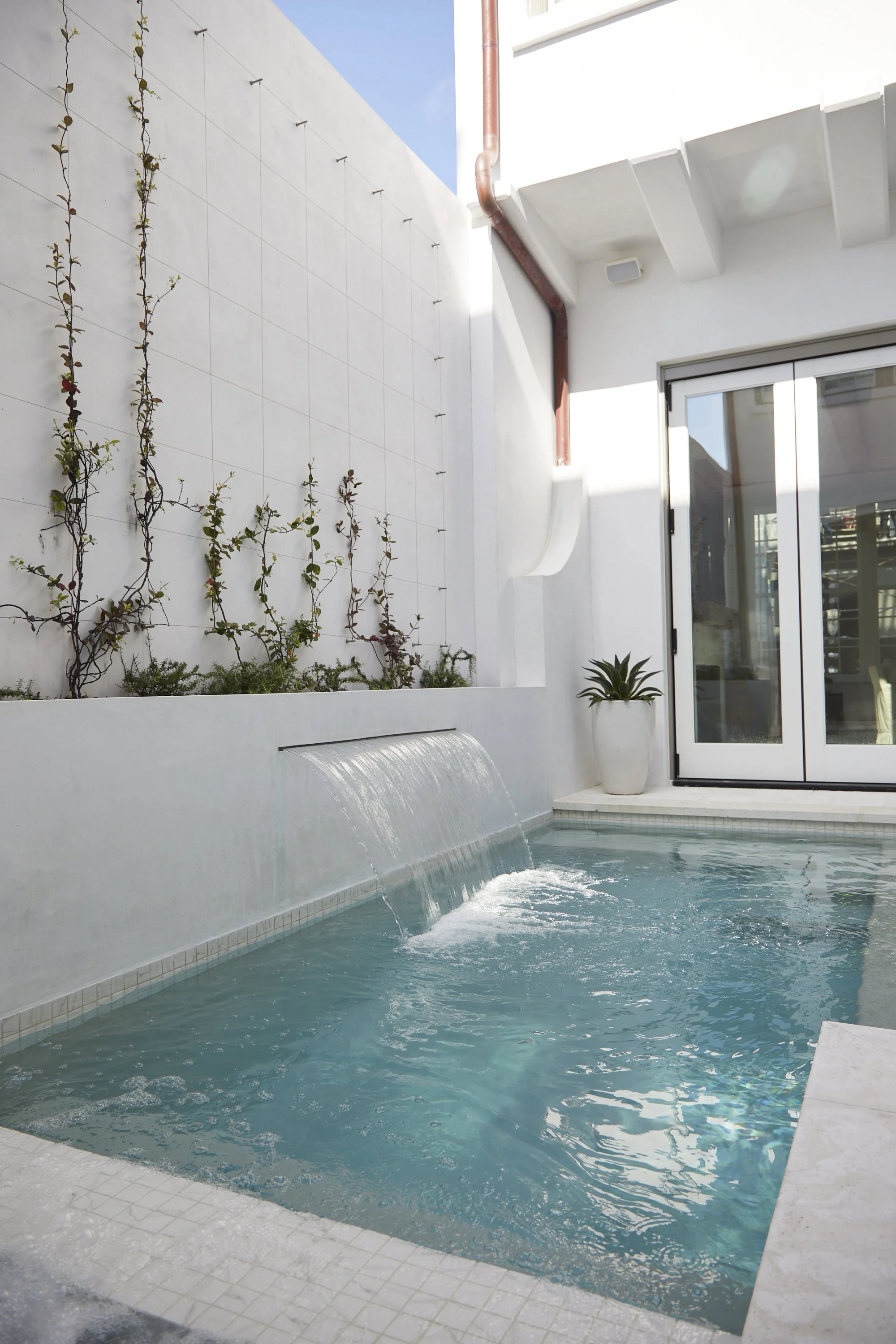 A modern backyard pool with a small waterfall feature, white walls, a potted plant, and sliding glass doors, with a clear blue sky above.