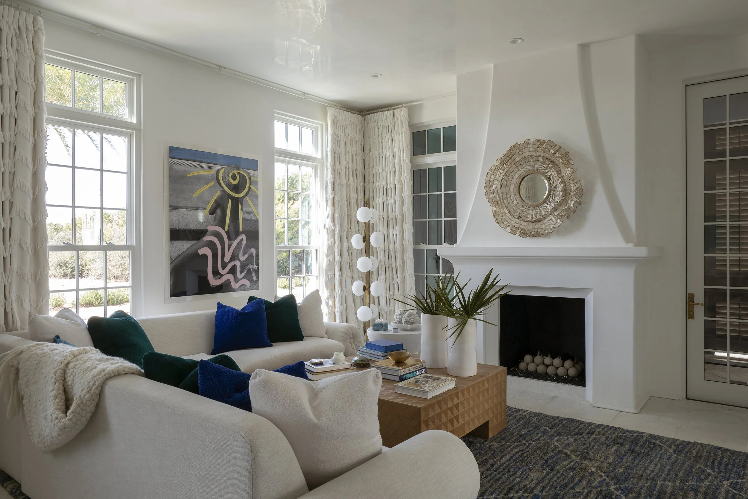 Living room with white sofa, blue and green pillows, a wooden coffee table, white rug, artwork on the wall, a white fireplace, large windows with white curtains, a plant, and a floor lamp with round white lights.