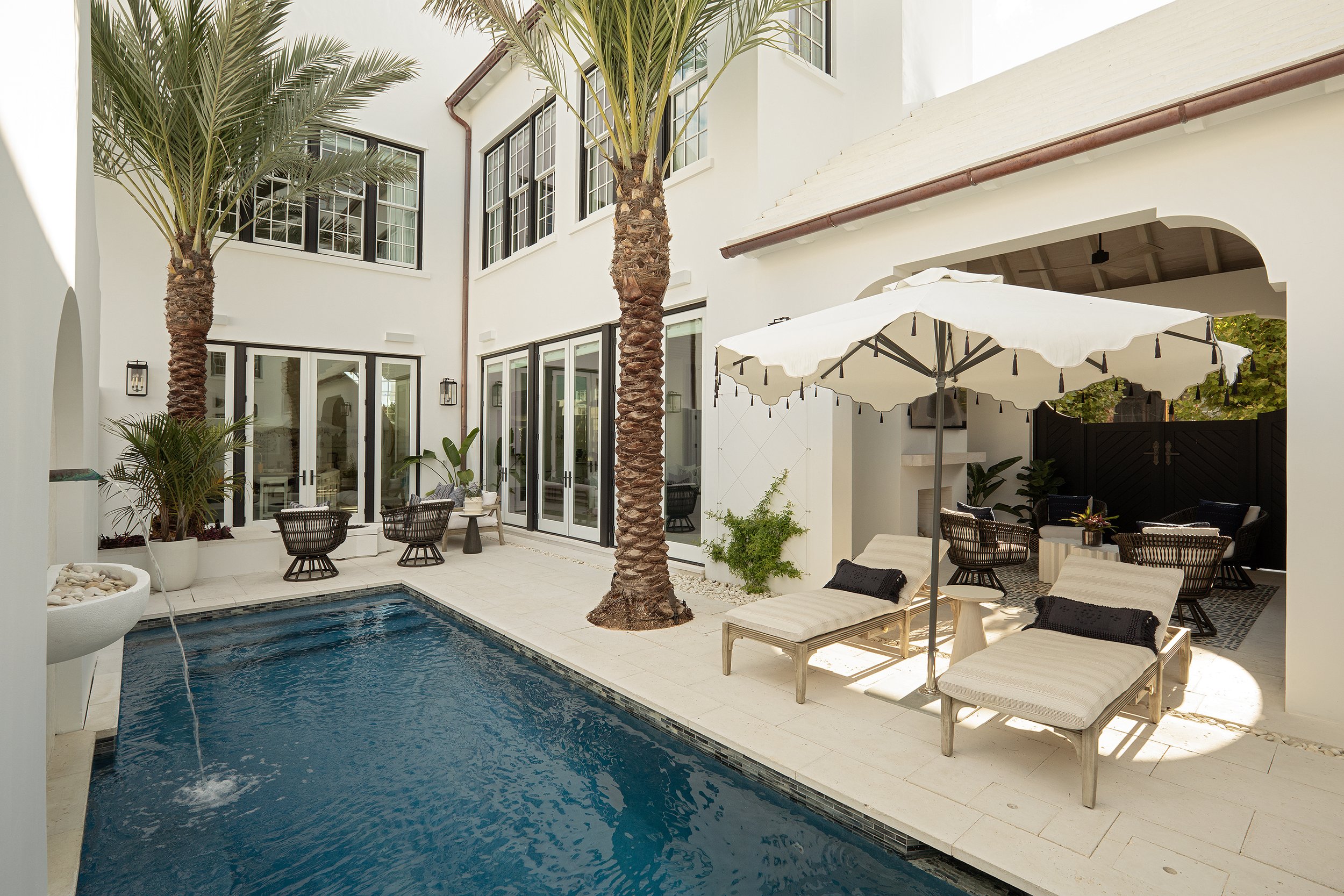 A private backyard with a swimming pool, lounge chairs with pillows, a large white umbrella, palm trees, potted plants, and outdoor seating areas, surrounded by white walls and large windows.