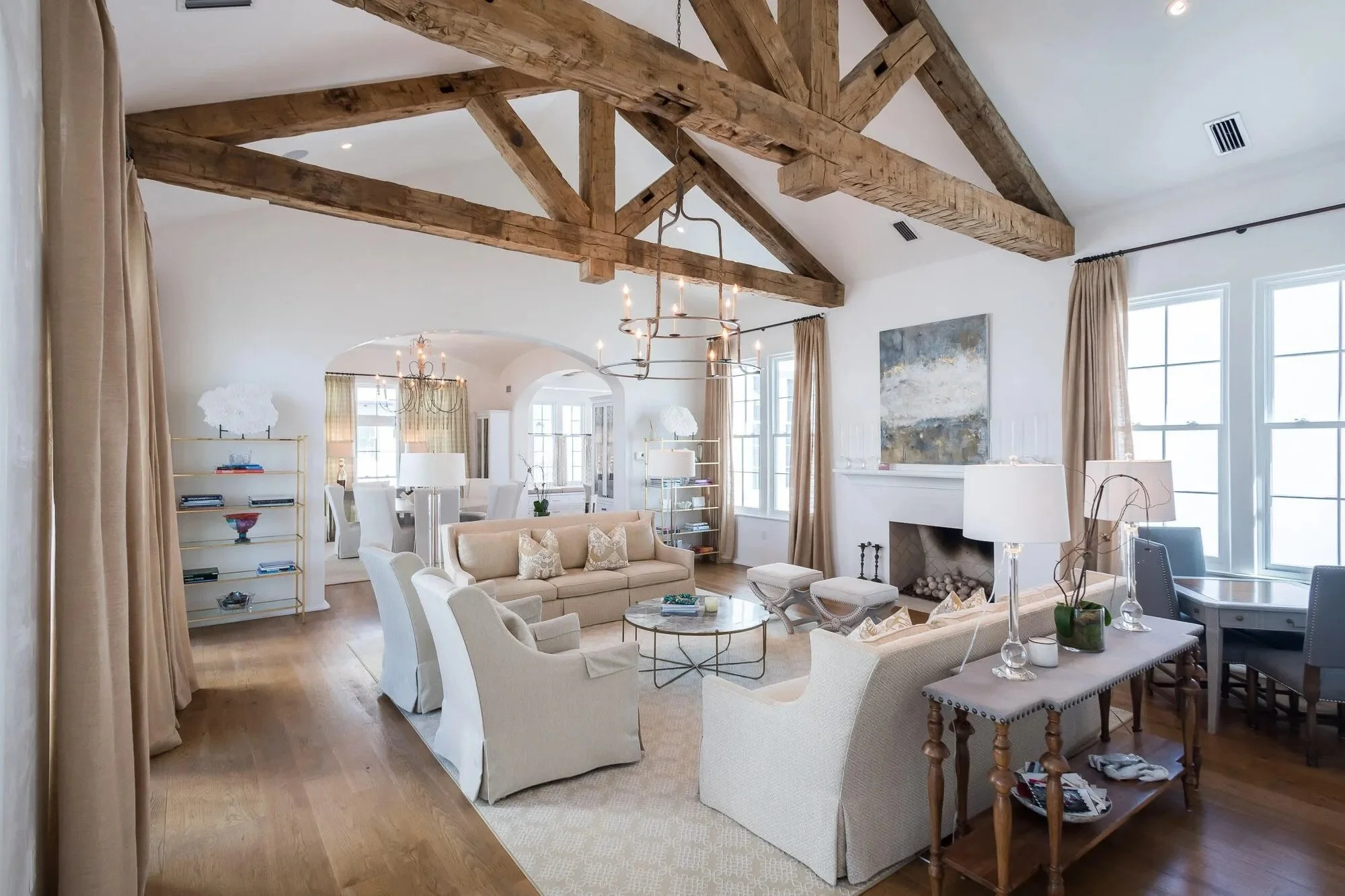 A spacious, well-lit living room with white walls, large windows, wooden ceiling beams, and modern furniture including cream sofas, an armchair, and a glass coffee table. There are lamps, artwork, and a fireplace, with an adjoining dining area visibl