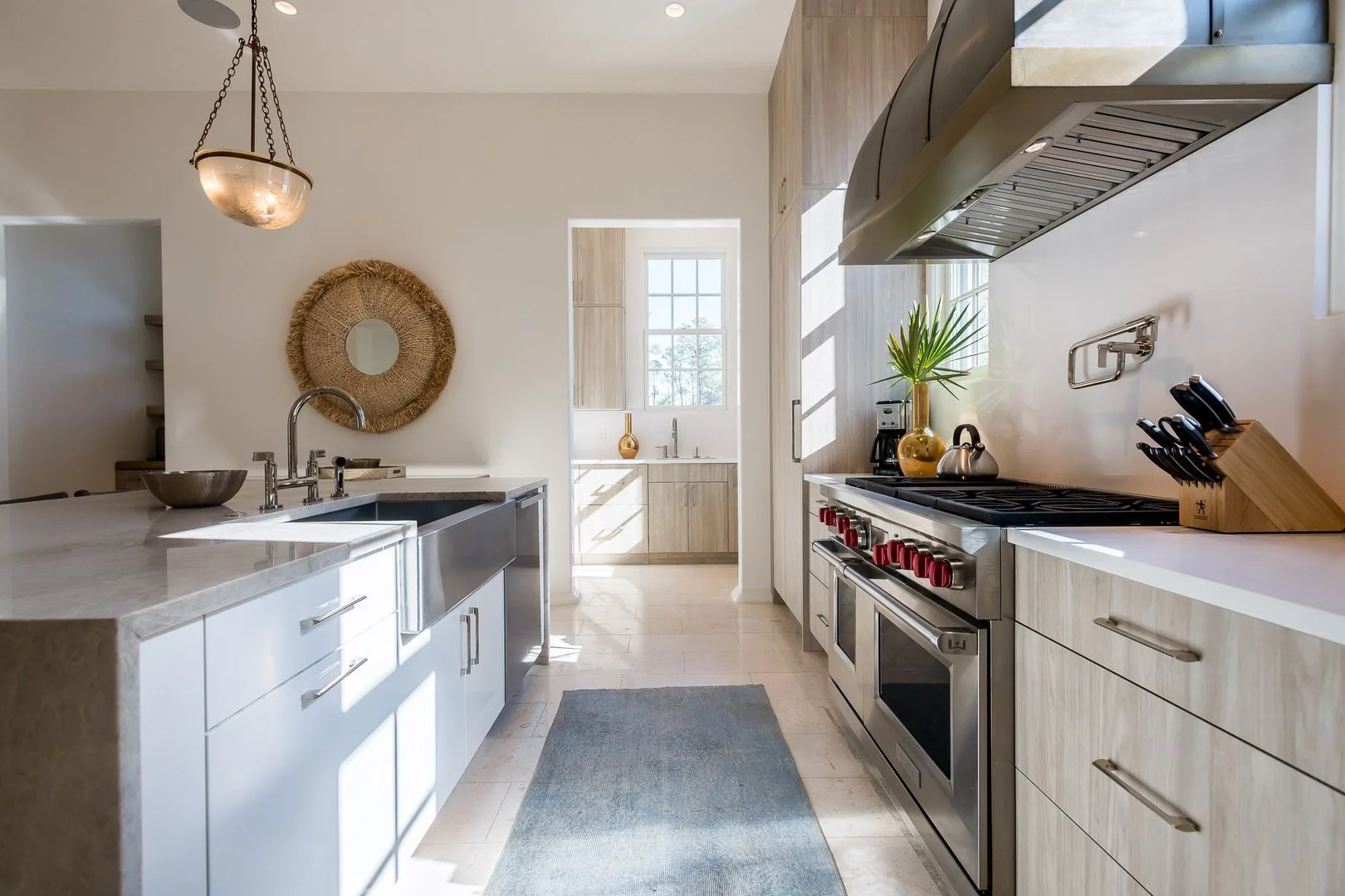 Modern kitchen with light wood cabinets, stainless steel appliances, a white countertop, and a blue rug on the tile floor. There is a pendant light and a round woven mirror on the wall.