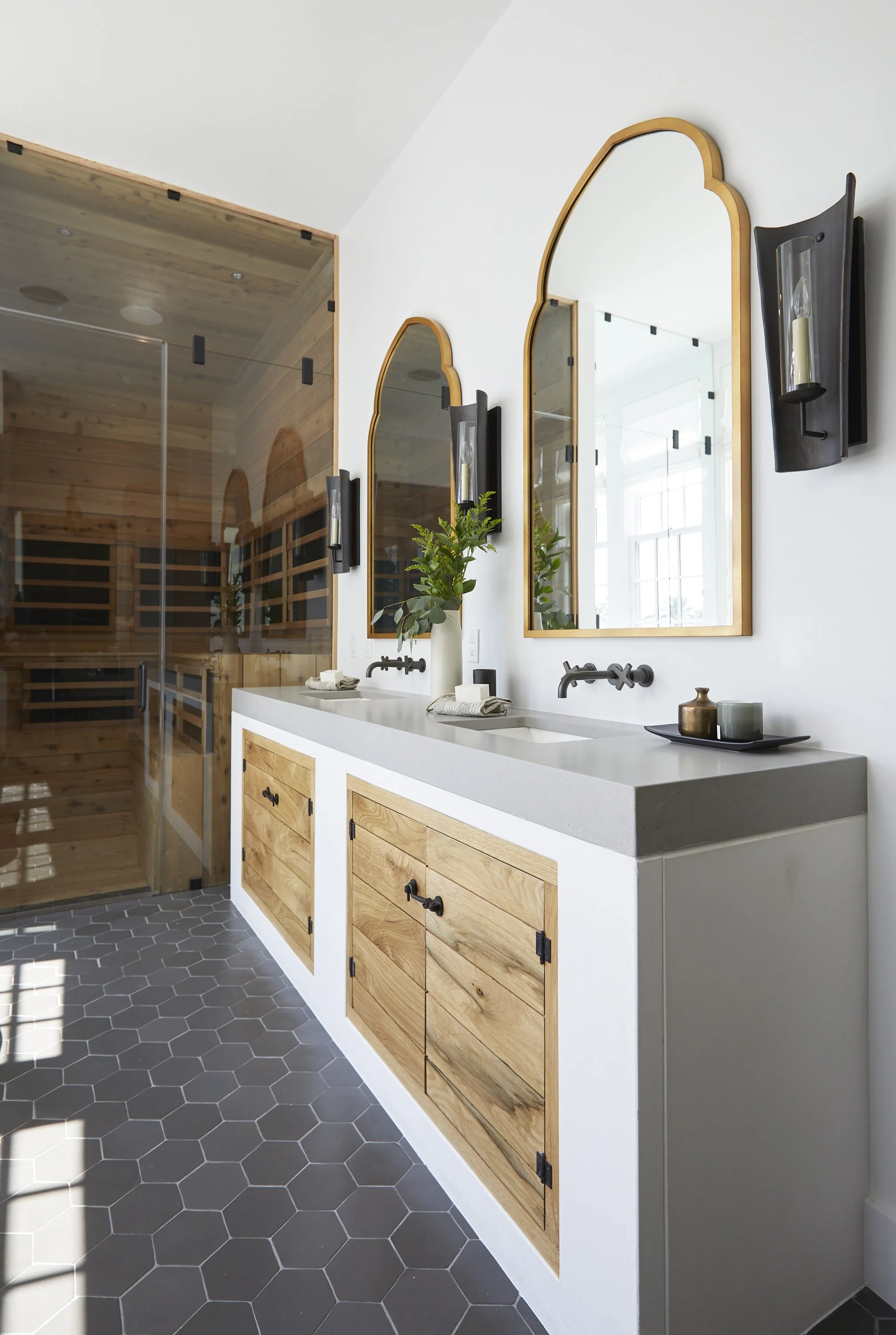 A modern bathroom with a double vanity featuring a white countertop, wooden cabinets, and two black wall-mounted faucets. Above the vanity are two arched mirrors with gold frames, flanked by black wall sconces with glass holders. A vase with greenery