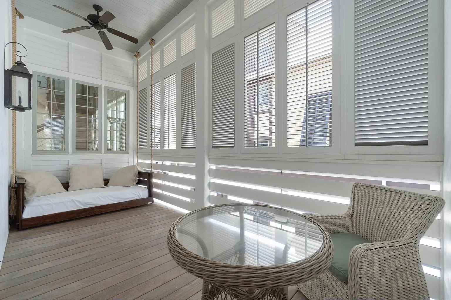 Sunroom with white wooden walls, multiple large windows with white blinds, a ceiling fan, wicker loveseat with cushions, round wicker table with glass top, and wicker chair.