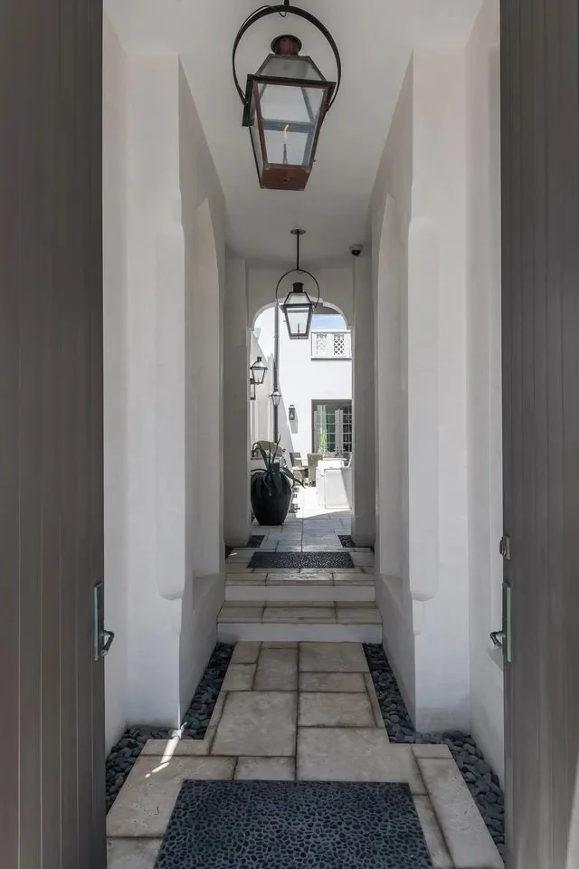 View of a white archway corridor leading to an outdoor patio with white furniture, black potted plant, and lantern-style lights hanging from the ceiling.