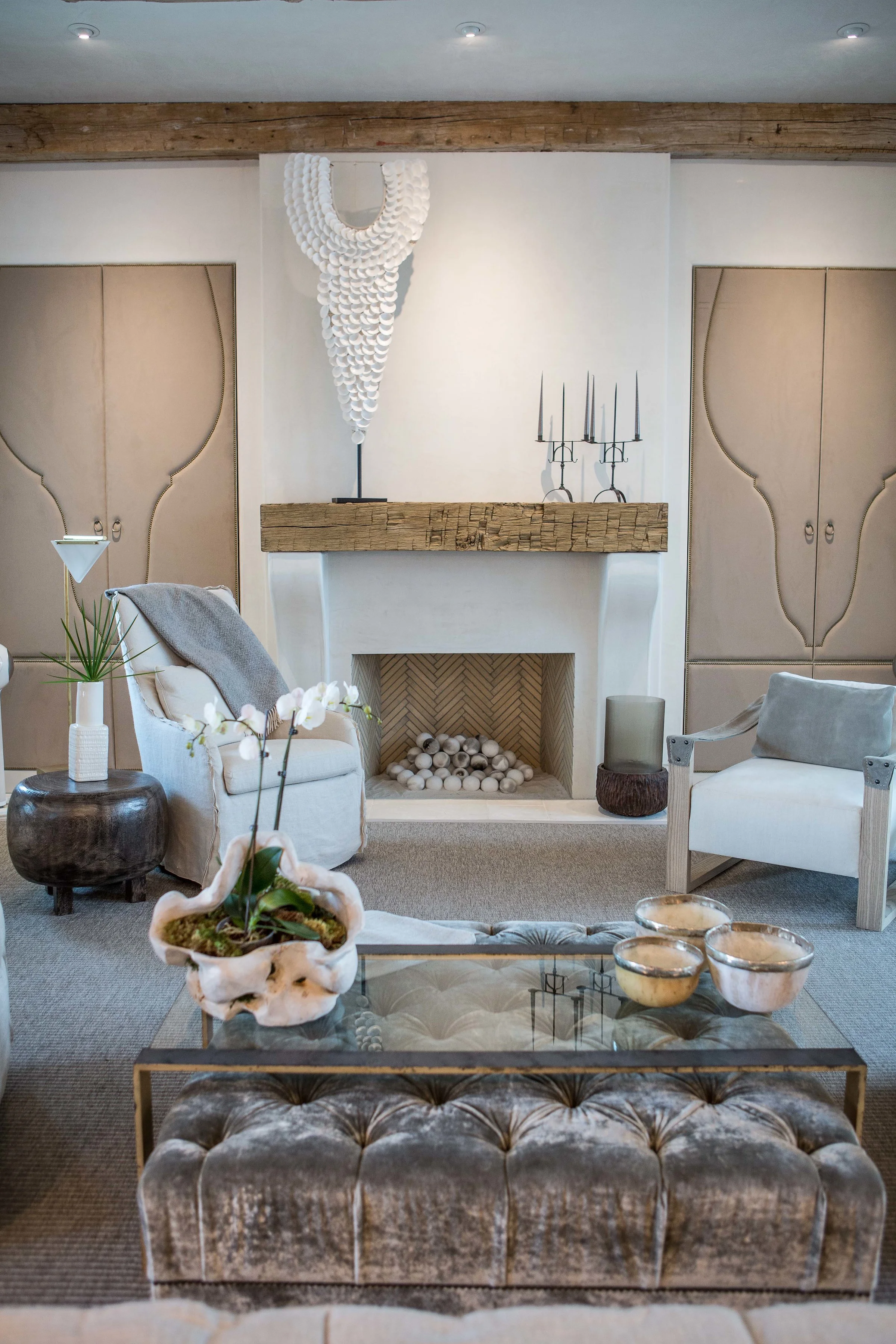 Living room with a white fireplace, beige sofas, a glass coffee table with bowls, and oxidized metallic side tables, decorated in a neutral modern style.