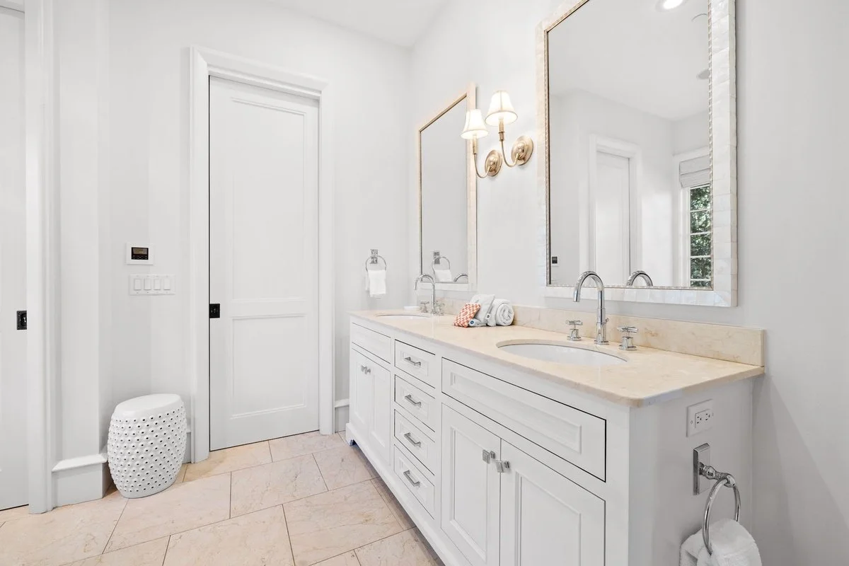 Bright white bathroom with double vanity, large mirror, wall-mounted lights, and a small white stool.
