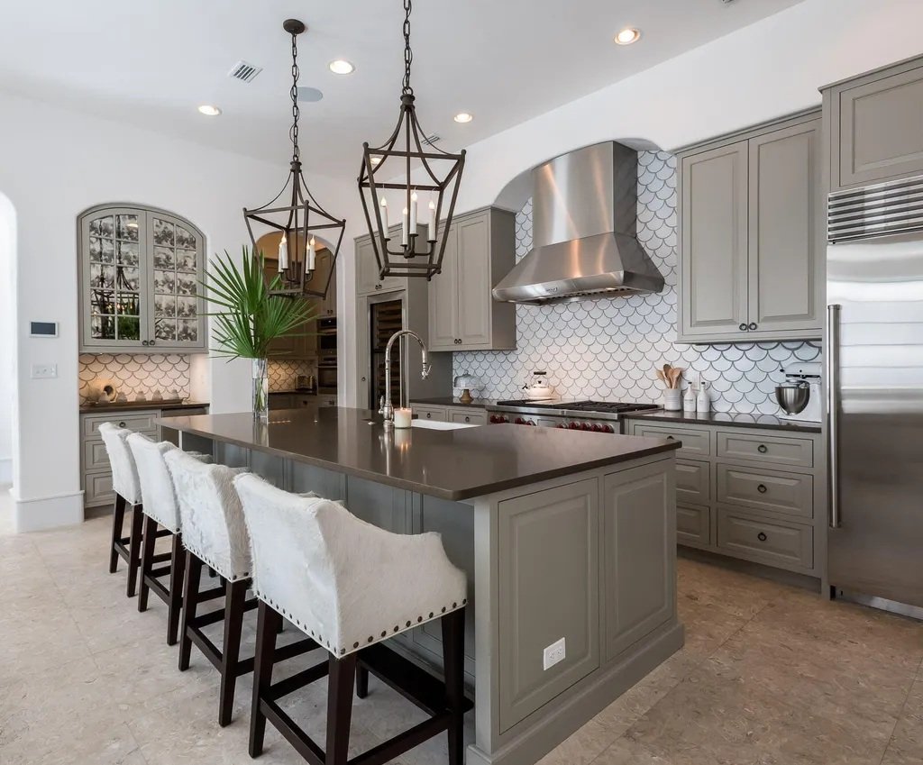 Modern kitchen with a large central island, gray cabinetry, stainless steel range hood, patterned white tile backsplash, and two black lantern pendant lights hanging over the island. Four white upholstered bar stools with dark wood legs are at the is