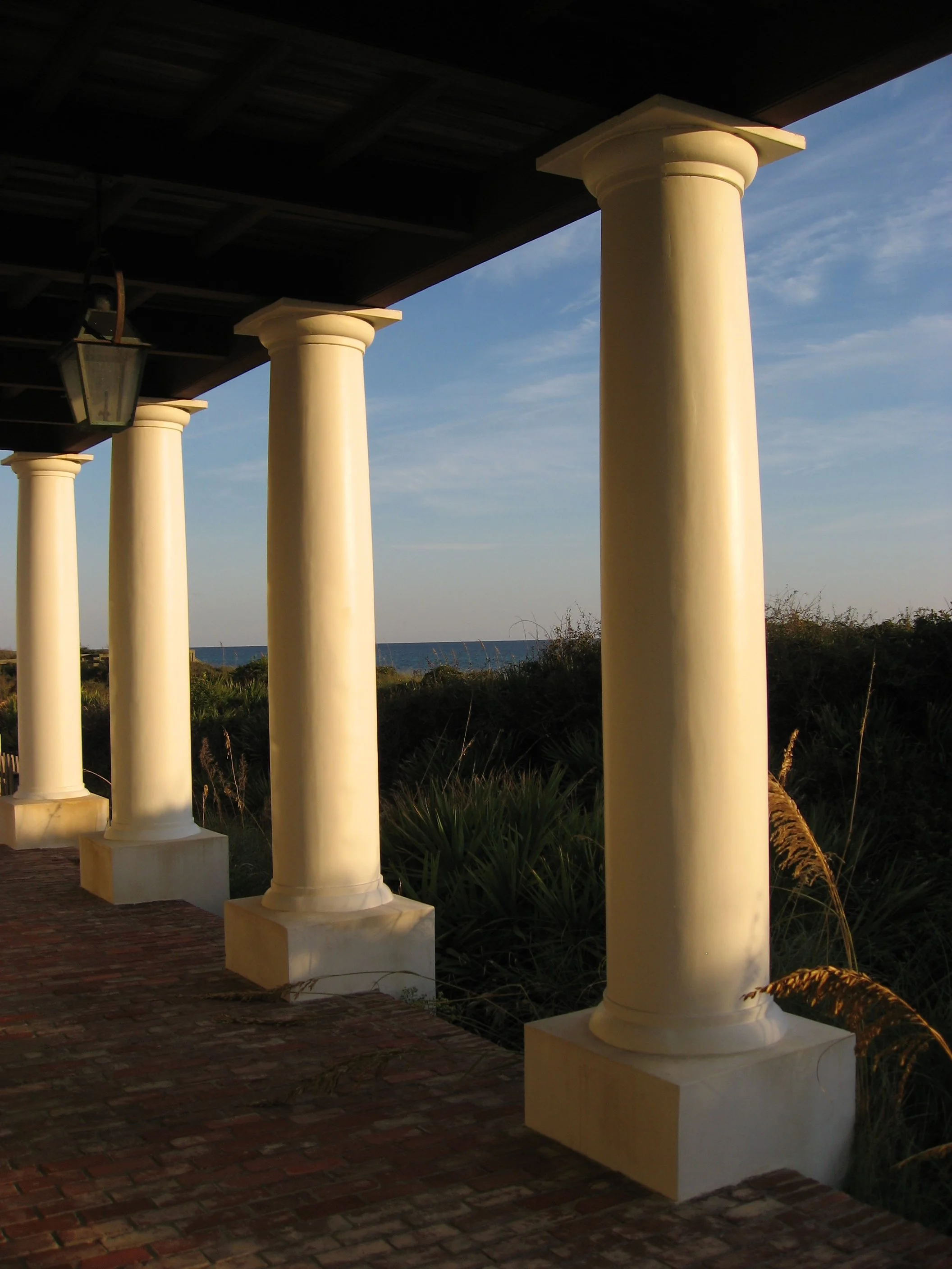 A view of a porch with white columns supporting a dark roof, overlooking a grassy area and the ocean in the distance during sunset or sunrise.