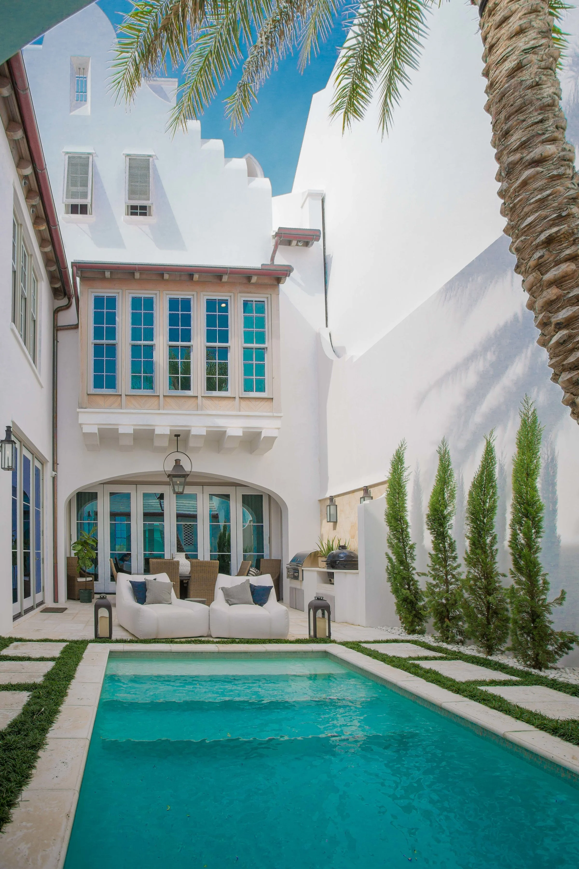 A backyard with a rectangular swimming pool, outdoor seating with two white chairs, a dining table with chairs, tropical plants, and a white multi-story house with large windows and a balcony, under a bright blue sky.