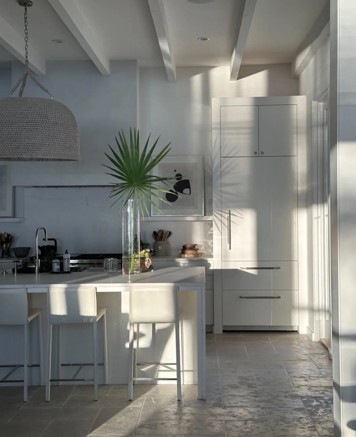 Modern kitchen with white cabinets, a kitchen island with four white bar stools, a large vase with a green palm leaf, and sunlight casting shadows on the wall.
