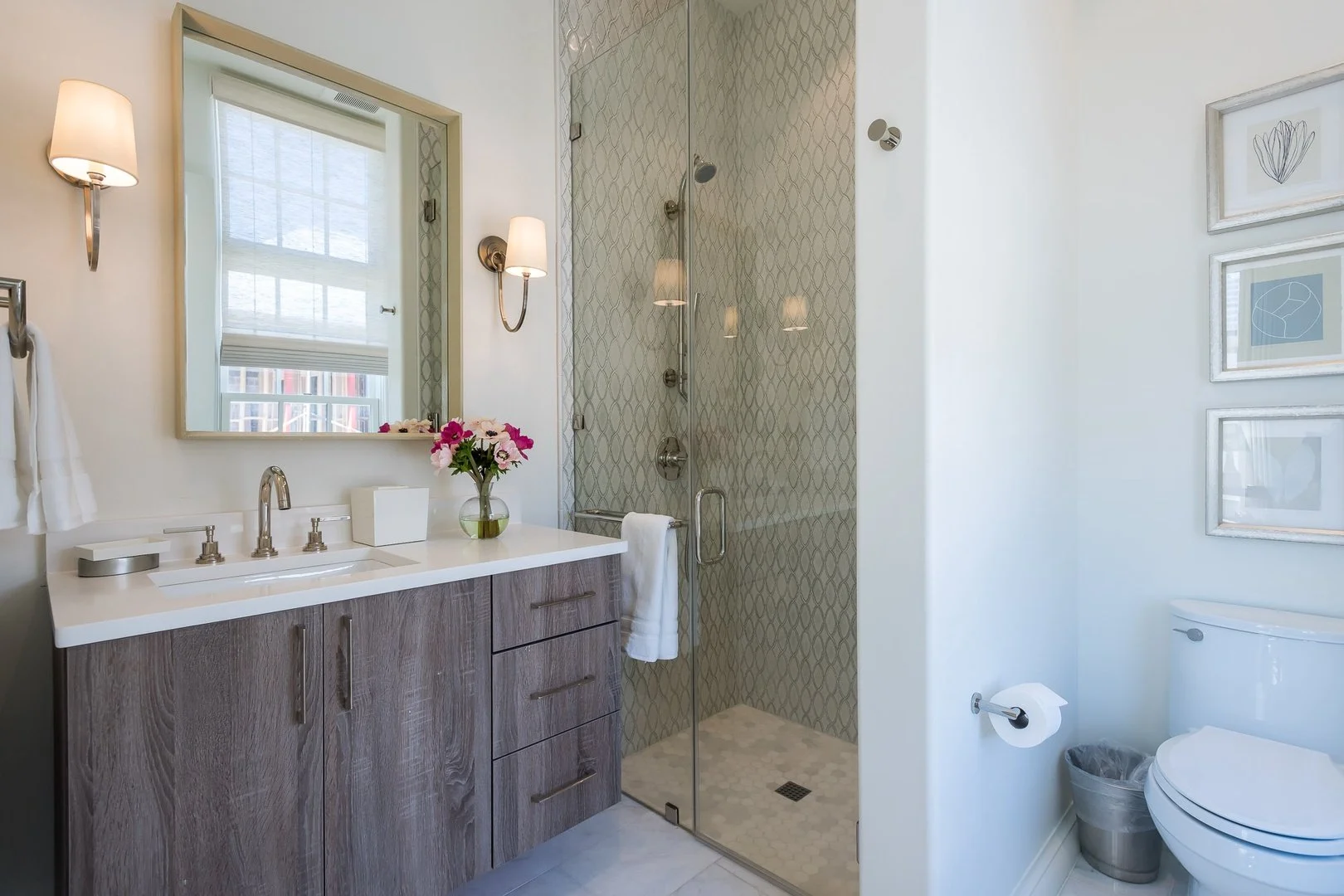 A modern bathroom with a wooden vanity, large mirror, and a flower vase. There is a glass-enclosed shower with patterned tiles, a white toilet, and framed artwork on the wall.