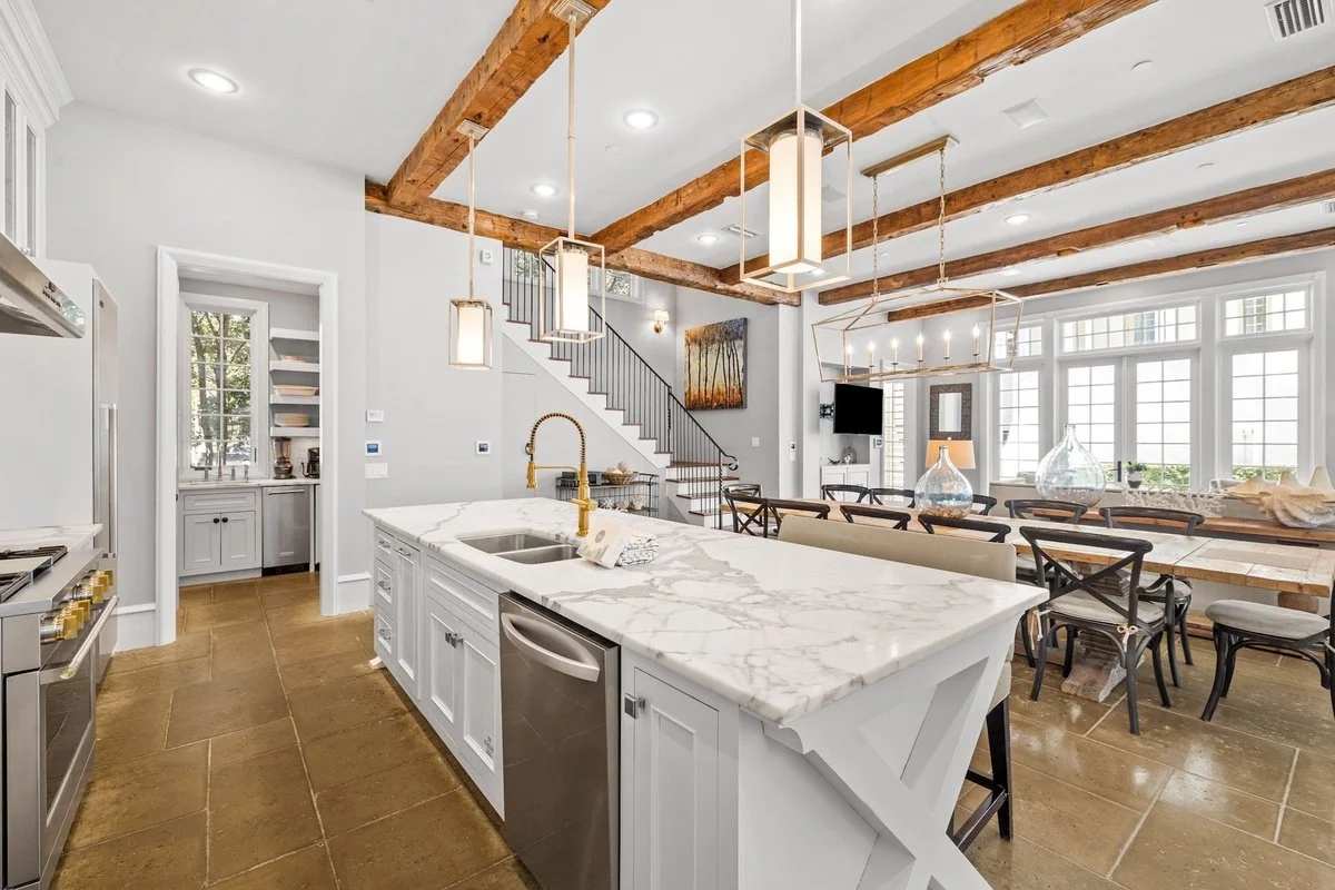 Open-concept kitchen and dining area with exposed wooden beams, white walls, and large windows, featuring a white marble kitchen island with a gold faucet, and a dining table with black chairs and glass vases.