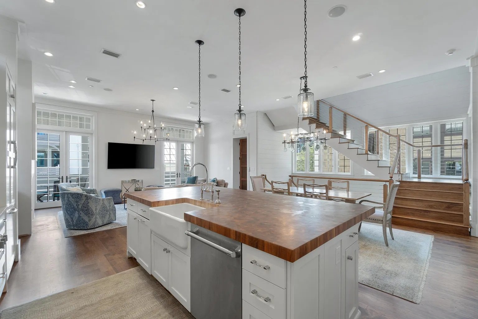 Open-concept living space with kitchen and living room, featuring a wooden island, white cabinetry, pendant lighting, hardwood floors, large windows, and stairs leading upstairs.