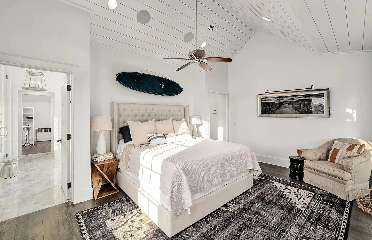 A bedroom with a high, white paneled ceiling, a beige upholstered bed with pillows, a wooden nightstand with books and a lamp, a dark area rug, a beige armchair with a pillow, a coffee table, a black and white wall art, and a surfboard mounted on the
