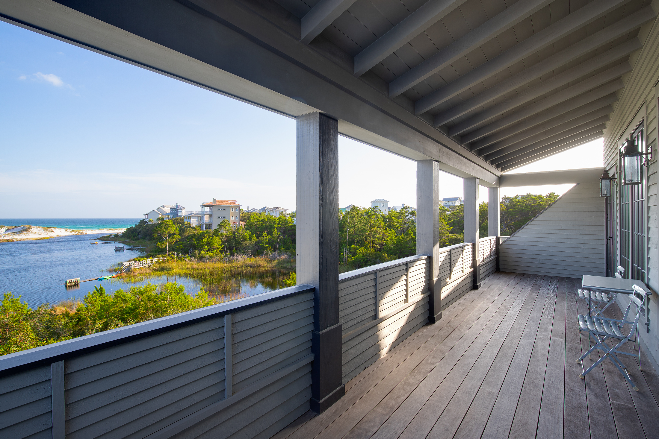 View from a balcony overlooking a body of water and houses in a coastal area, with a wooden deck and two metal chairs.