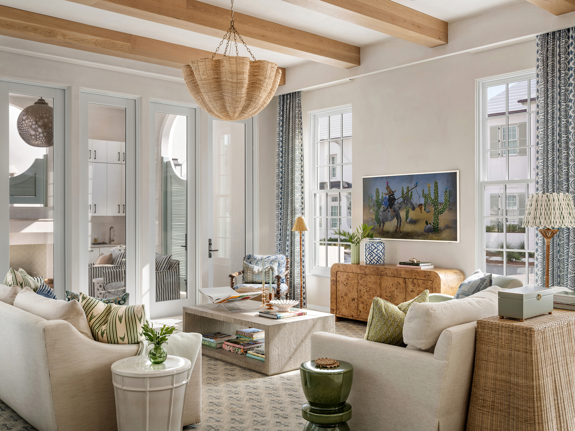 Bright living room with white sofas, a marble coffee table, and a wooden sideboard. Large windows with patterned curtains, and a flat-screen TV on the wall. Light wood ceiling beams and a woven chandelier.