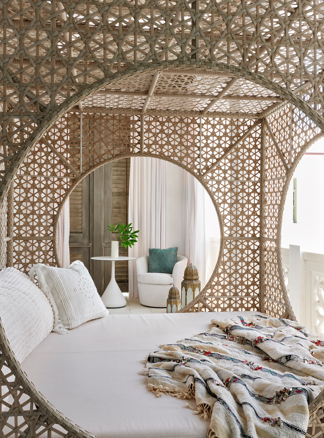 A bedroom with a round wicker canopy bed and white bedding. There are white pillows on the bed, a patterned throw blanket, and a cozy corner with a white armchair, green pillow, small white side table with a plant, and decorative lanterns.