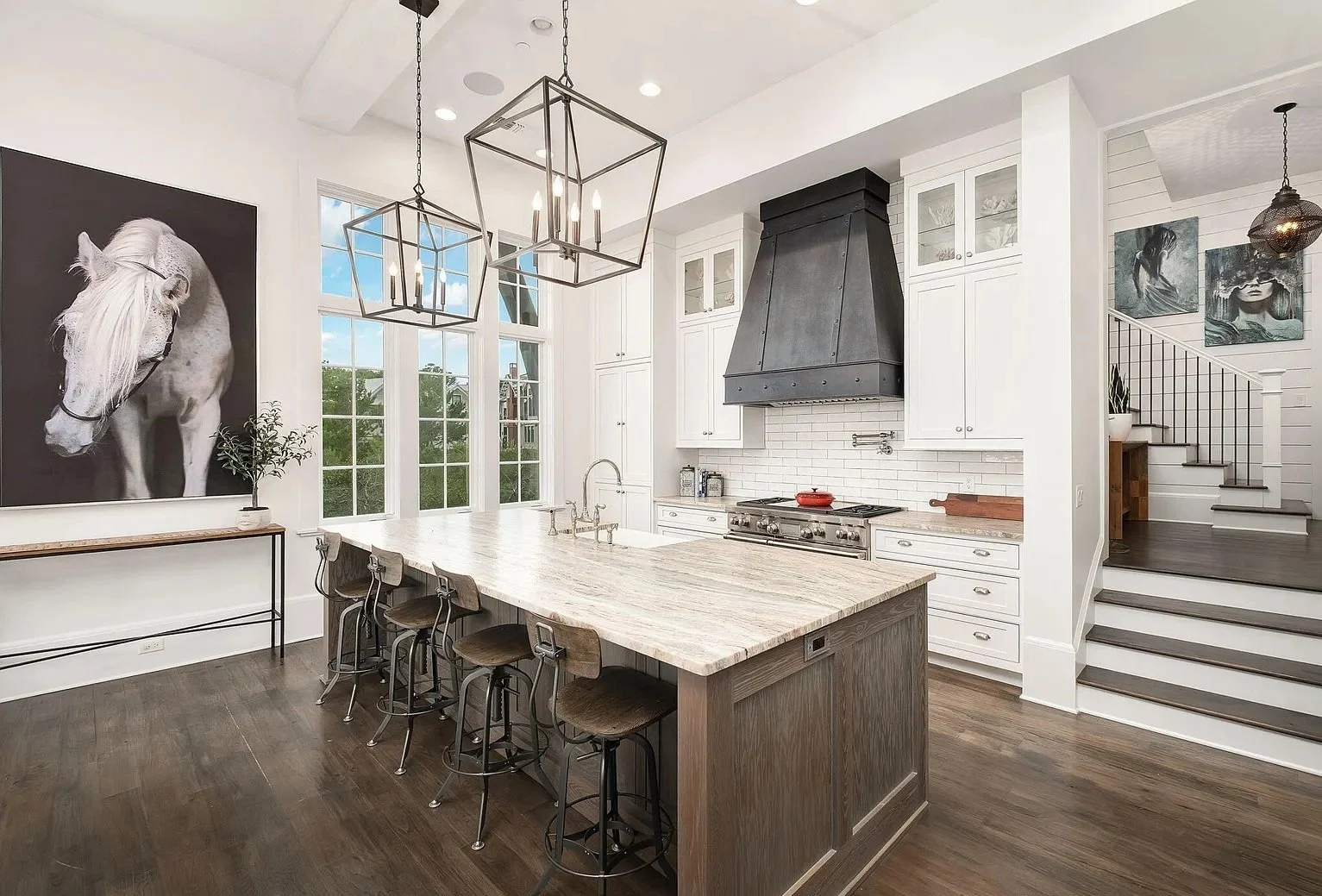 Modern kitchen with white cabinets, a large wooden island, black oven hood, and artwork including a large horse portrait and abstract images, with dark hardwood flooring.