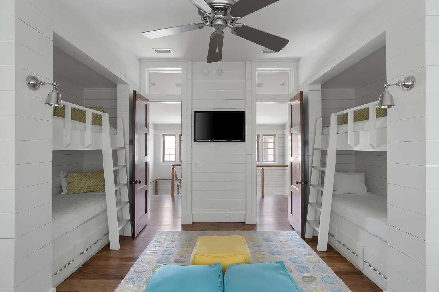 A bedroom with two sets of bunk beds built into the walls, each with white frames, green and white bedding, and ladders, mirrored closet doors, a wall-mounted TV, a ceiling fan with dark blades, a colorful area rug, a yellow ottoman, and blue pillows