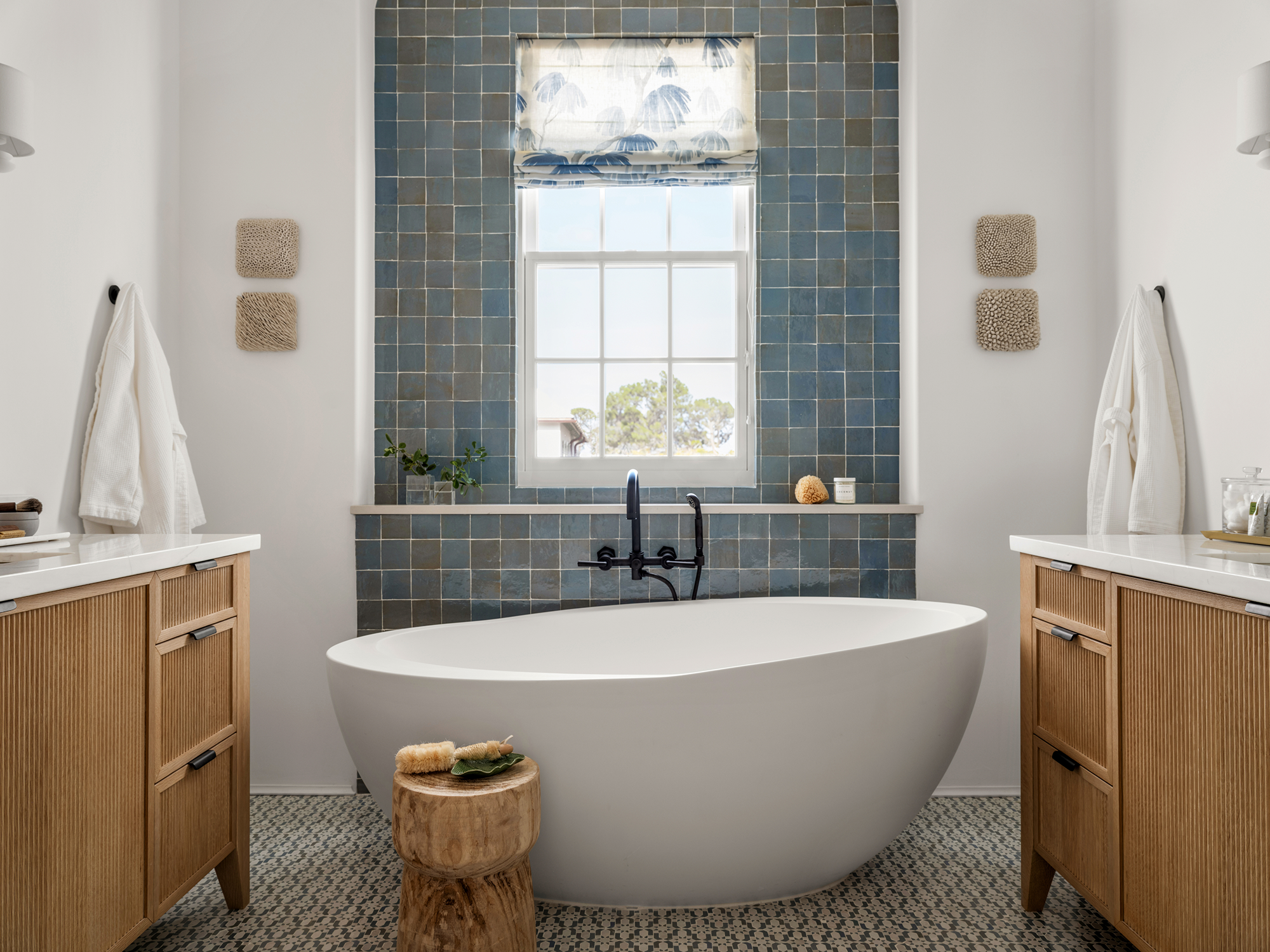 Modern bathroom with white freestanding bathtub, blue tiled accent wall, wooden vanity cabinets with white countertops, and a window above the tub, decorated with plants and small decor items.